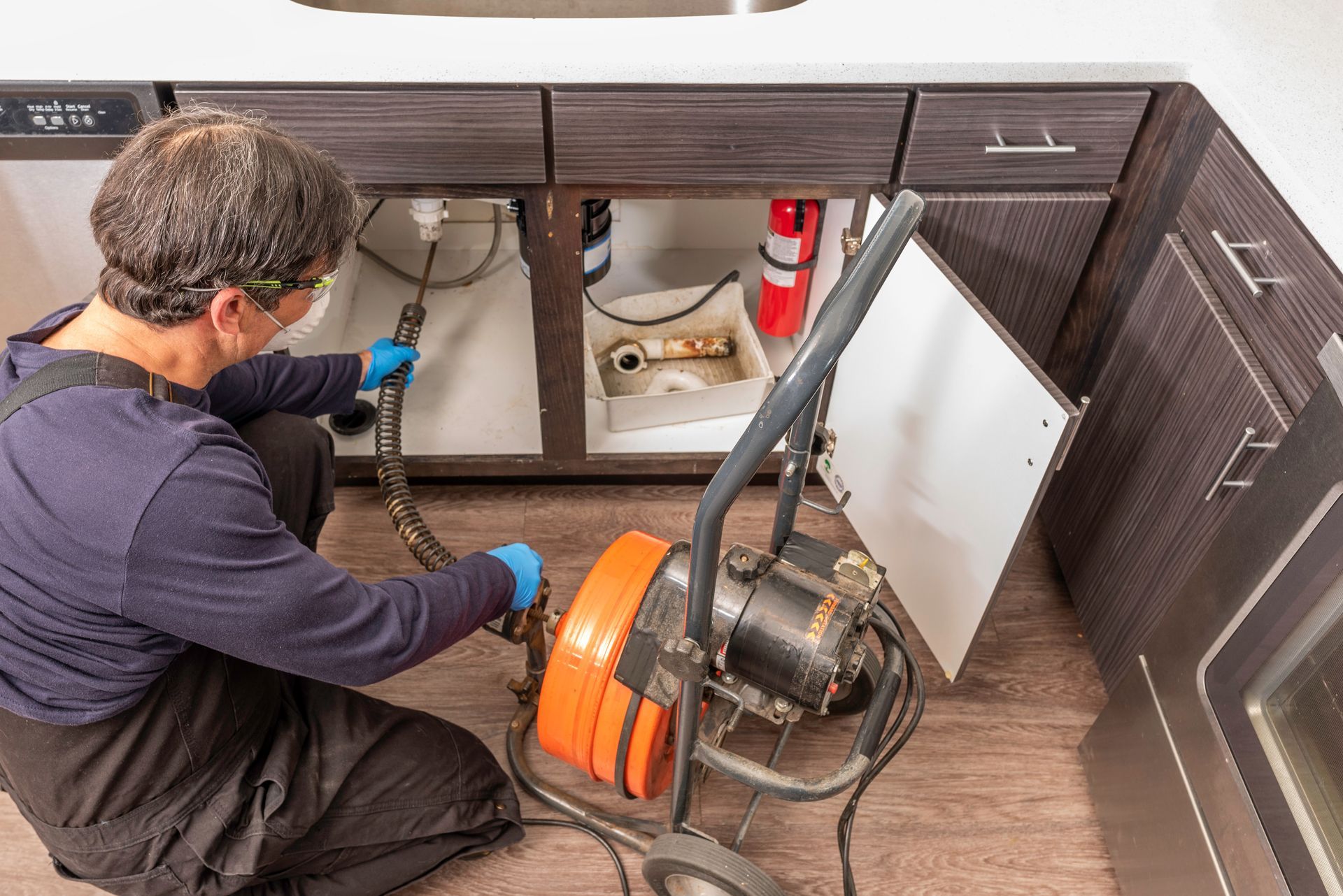 A plumber in blue gloves uses a professional orange drain auger to clear a clog under a kitchen sink.