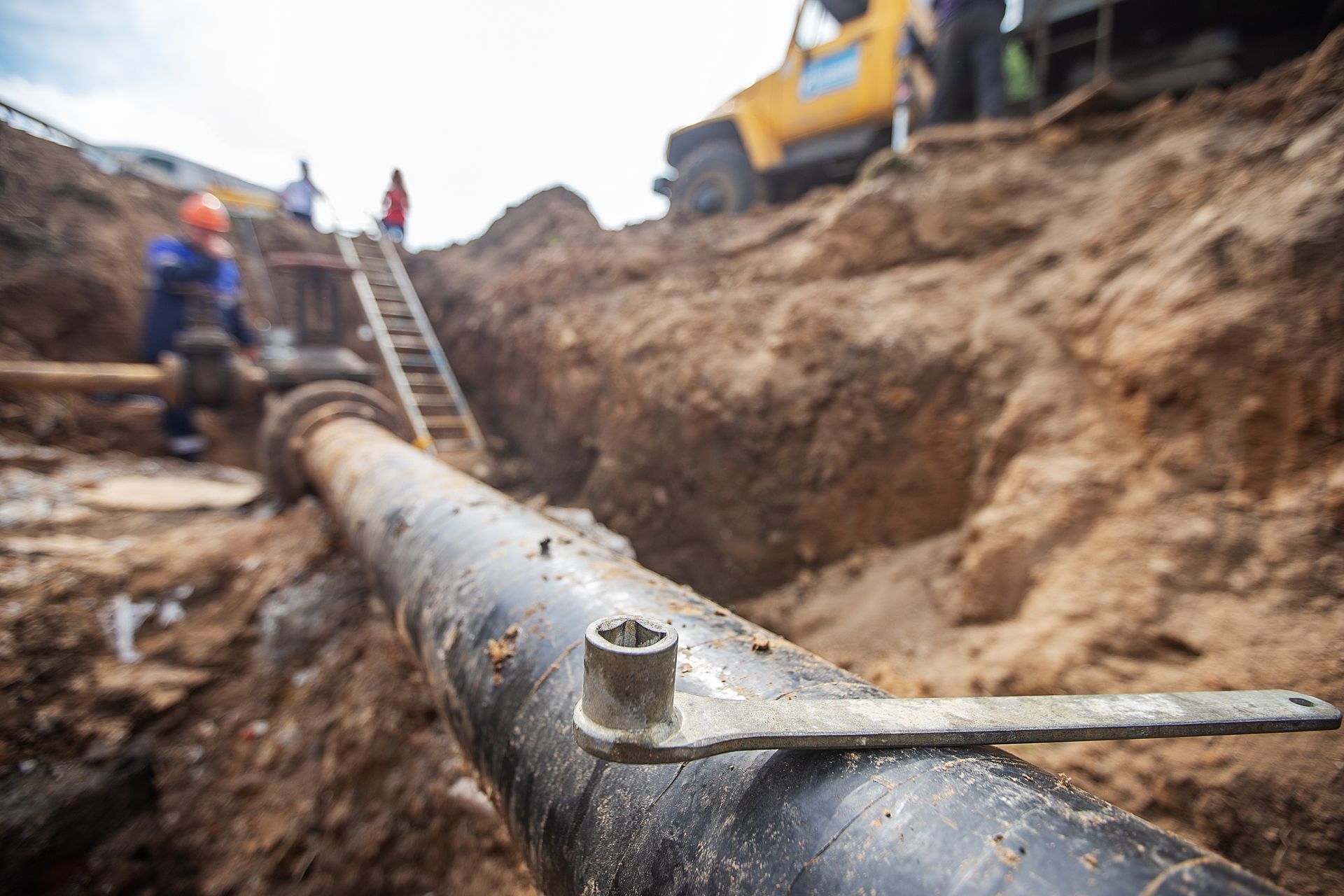 A wrench rests on a large pipe in an open trench, with construction workers and a yellow vehicle in the background.
