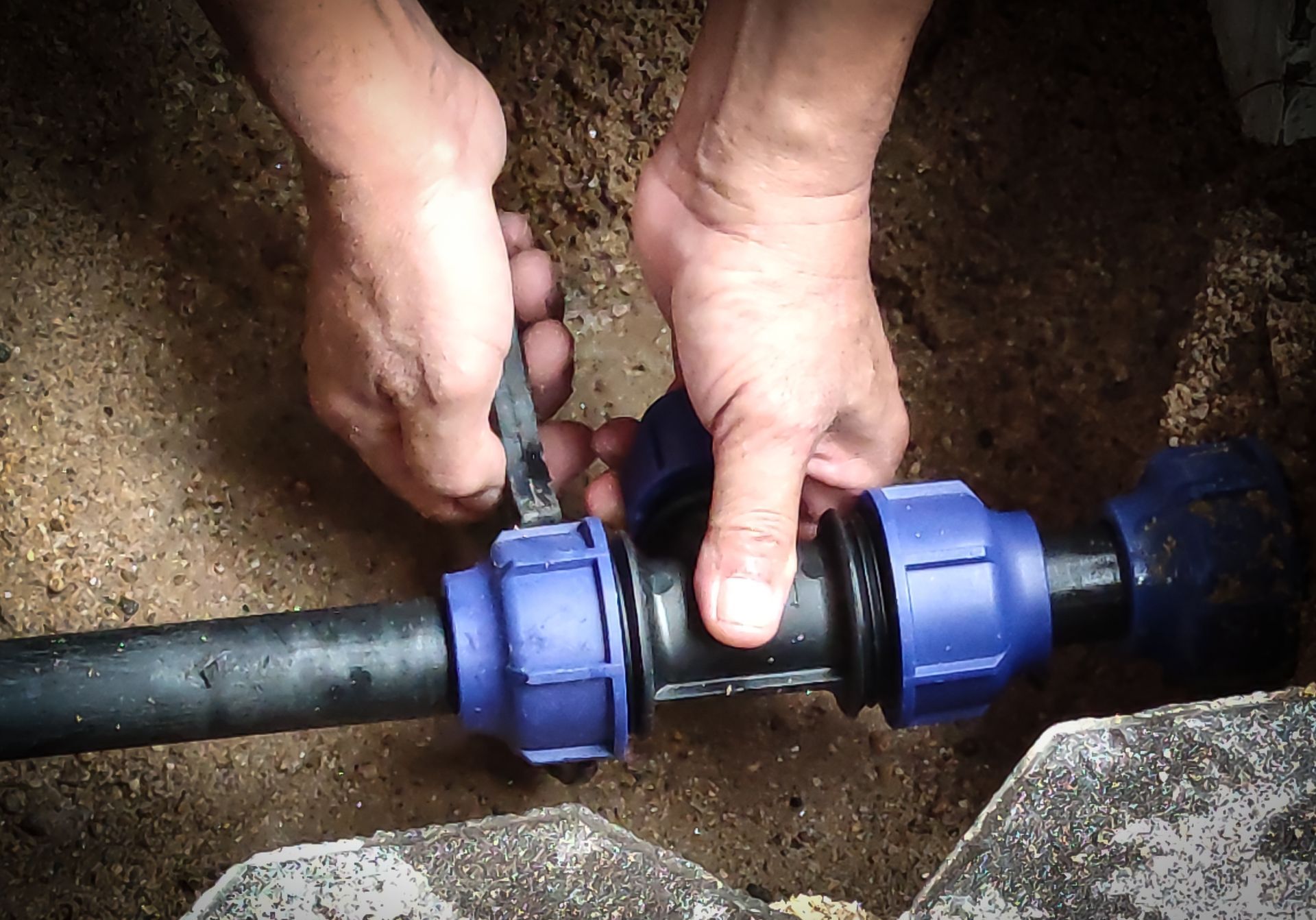 A person’s hands tighten a blue compression coupling onto a black pipe in a dirt trench.