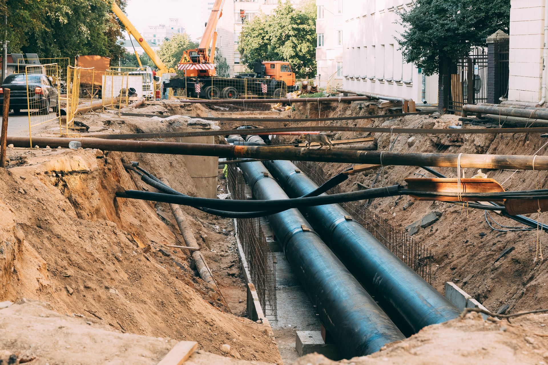A trench construction site with large, dark pipes running through the center, overseen by a crane in the background.