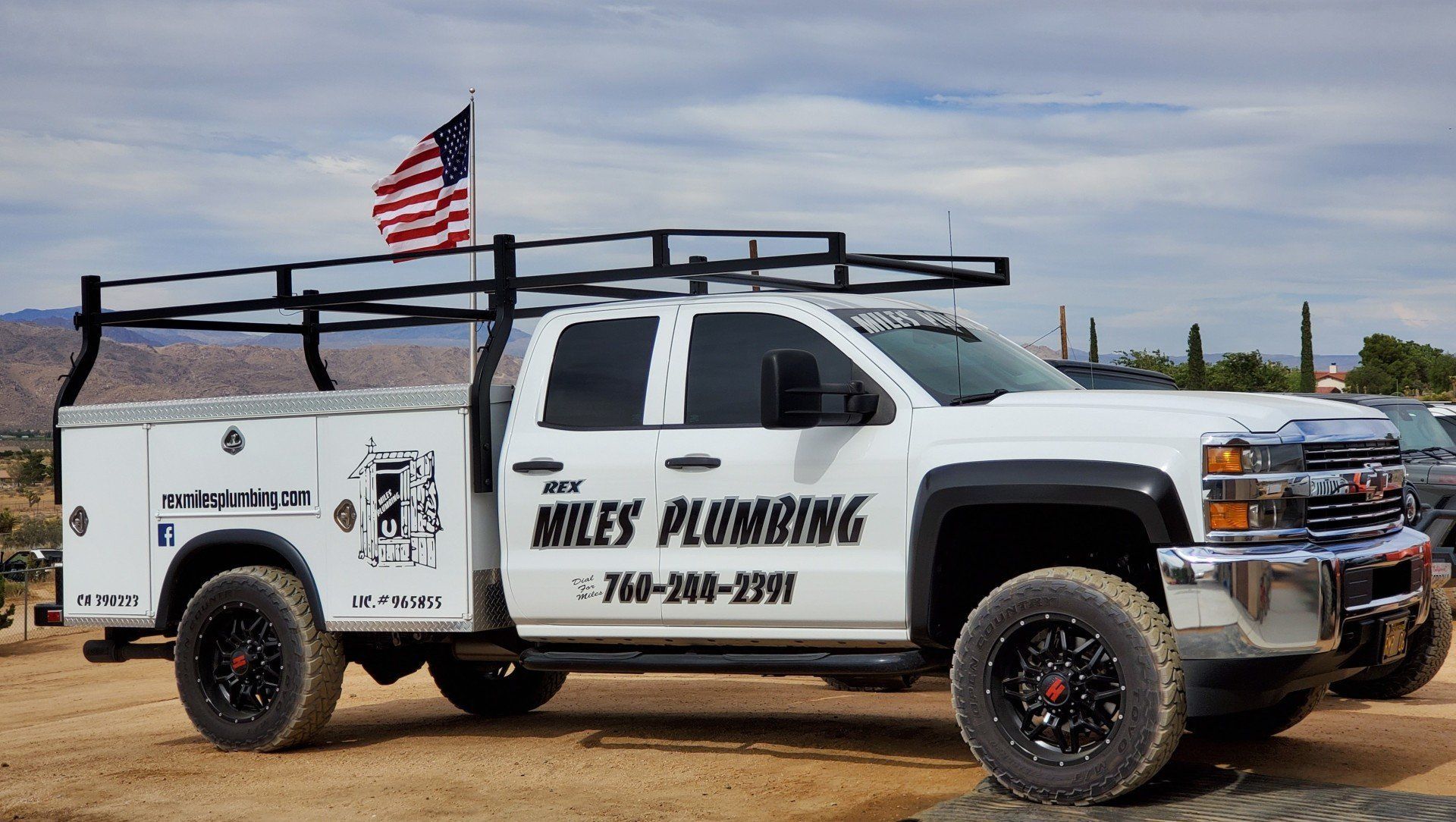 A white Miles Plumbing service truck with an American flag attached to a roof rack, parked on a dirt lot.