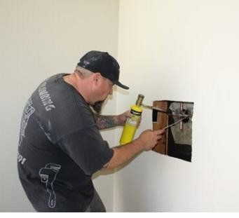 A worker uses a yellow handheld torch to solder copper pipes inside an open section of a white wall.