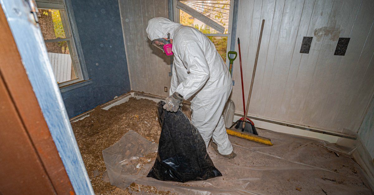 A person in a white protective suit placing debris into a black bag inside a room with tools.