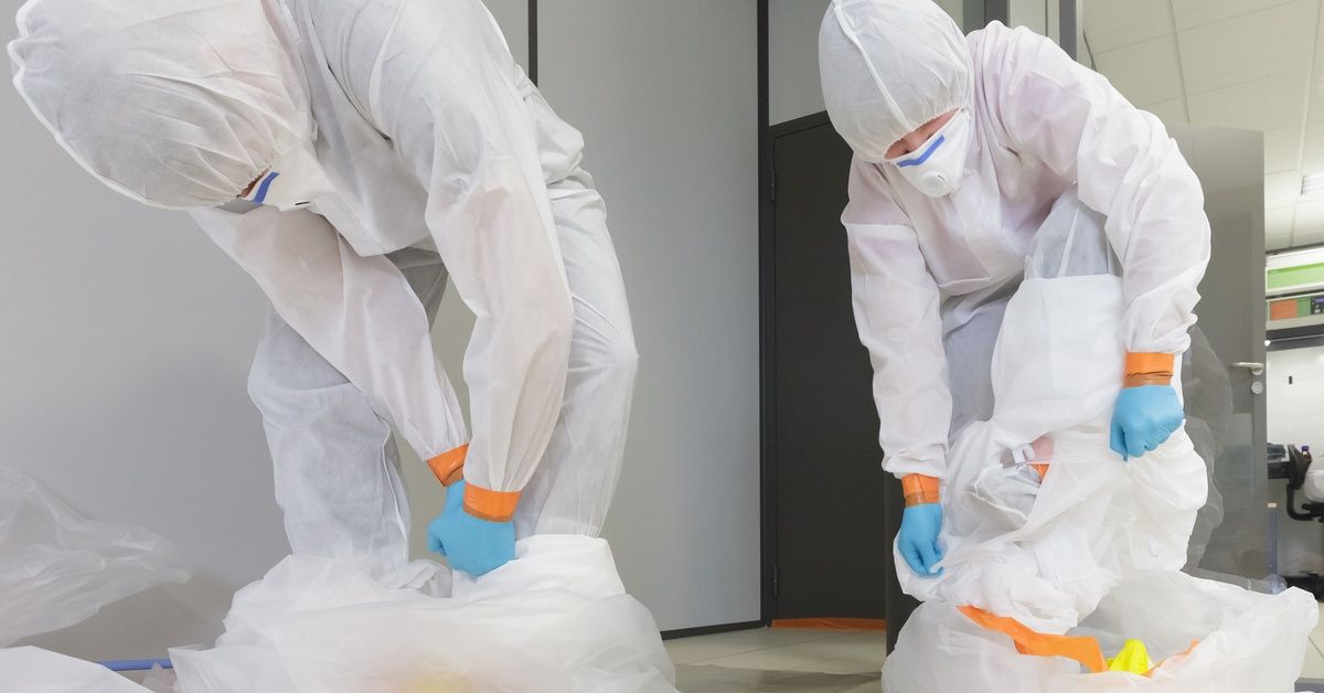 Two people wearing type 5/6 hazmat suits wrap up two plastic disposal bags in a room.