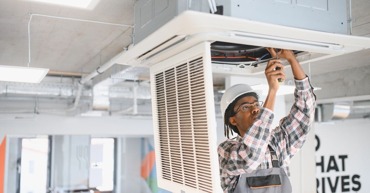 A technician has a tool in his hands and is reaching upwards within a large AC unit on the ceiling.