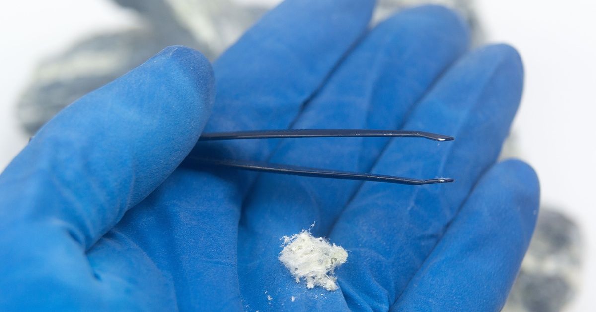 A close-up of a hand wearing a blue glove and holding a pair of tweezers and a pinch of asbestos.