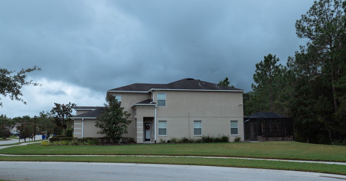 A large, beige house is sitting on a large, green lawn, alongside a road with stormy clouds.