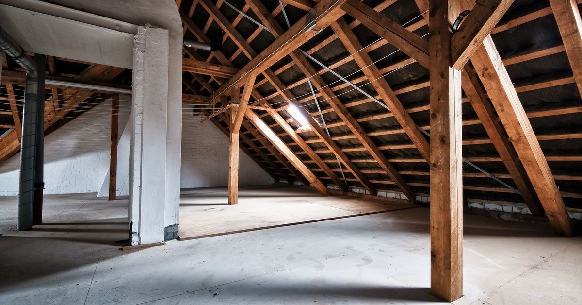 A large empty attic with exposed wooden beams, a concrete floor, and white brick walls.
