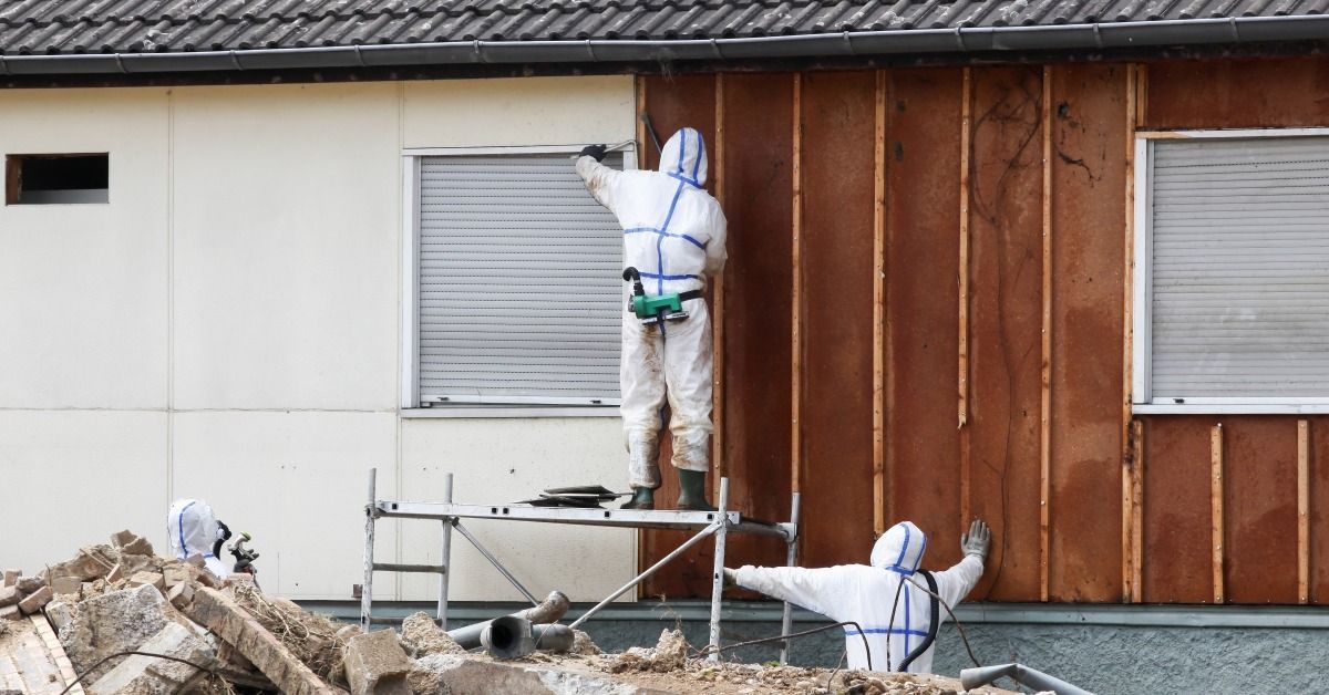 Workers in protective suits remove exterior wall materials from a home during asbestos abatement.