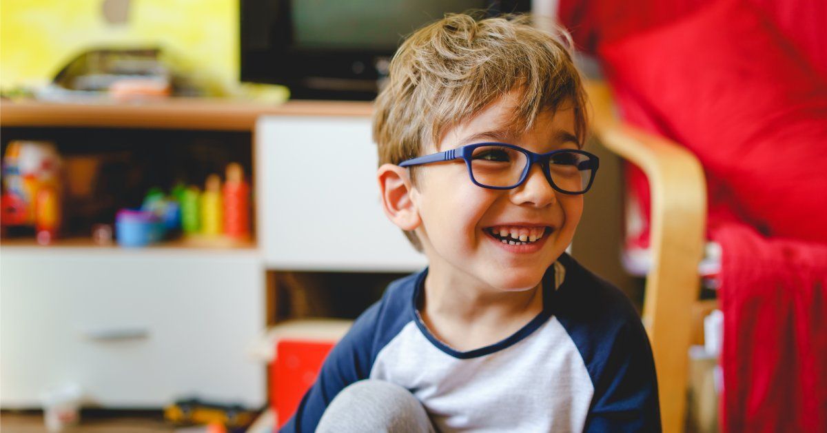 A young boy wearing glasses smiles while playing on a wooden floor.