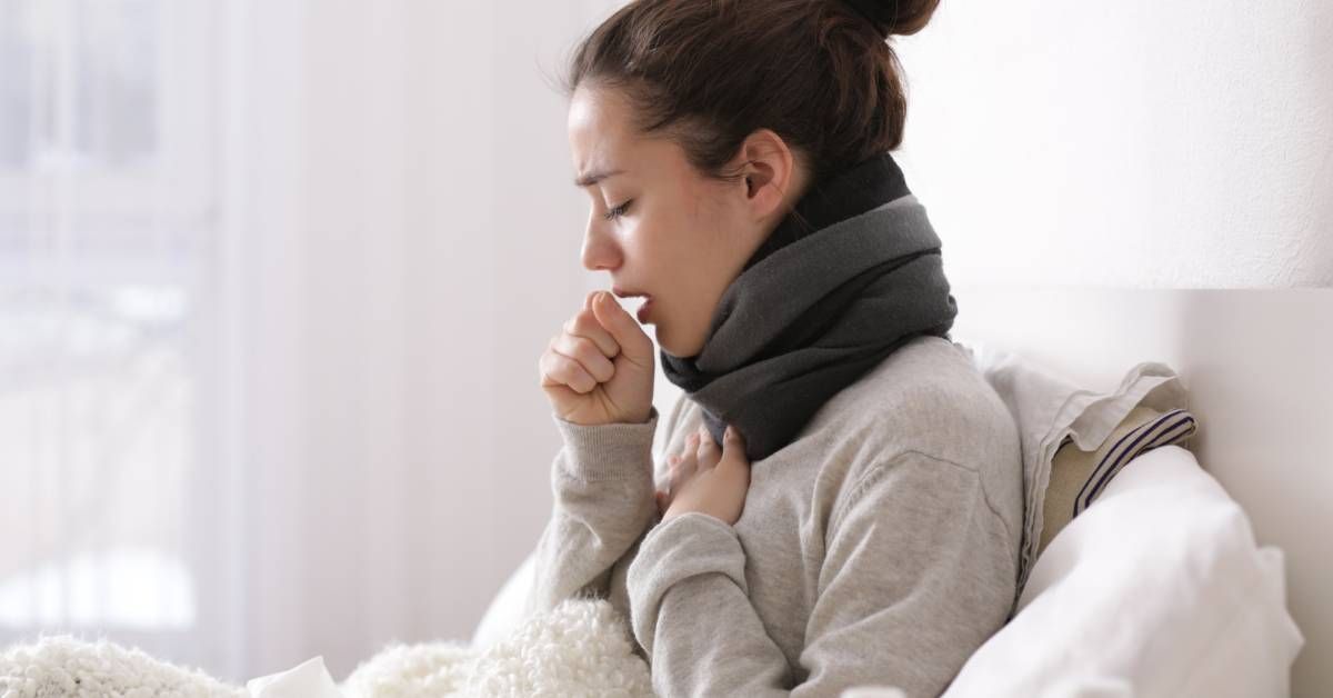 A young woman sits in bed and coughs into her hands. She has a blanket on her lap and a scarf.