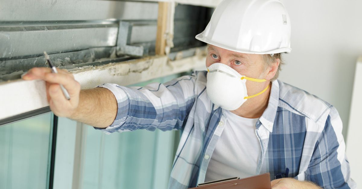 A man wearing a flannel, hard hat, and face mask inspects a home. He's holding a clipboard and pen.