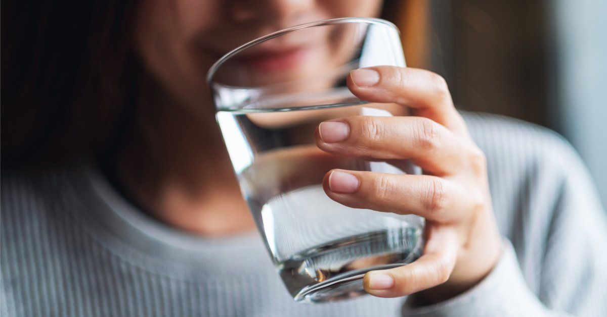 A woman in a gray sweater holding a glass of water up to her mouth. She has a slight smile.