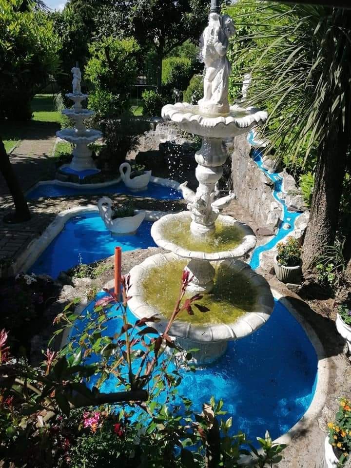 Giardino con fontane a più livelli, acqua azzurra e statue bianche, circondato dal verde.