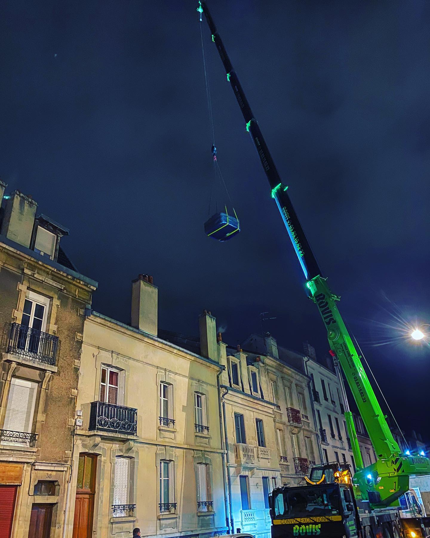 Une grue verte soulève une boîte dans les airs devant un bâtiment la nuit.