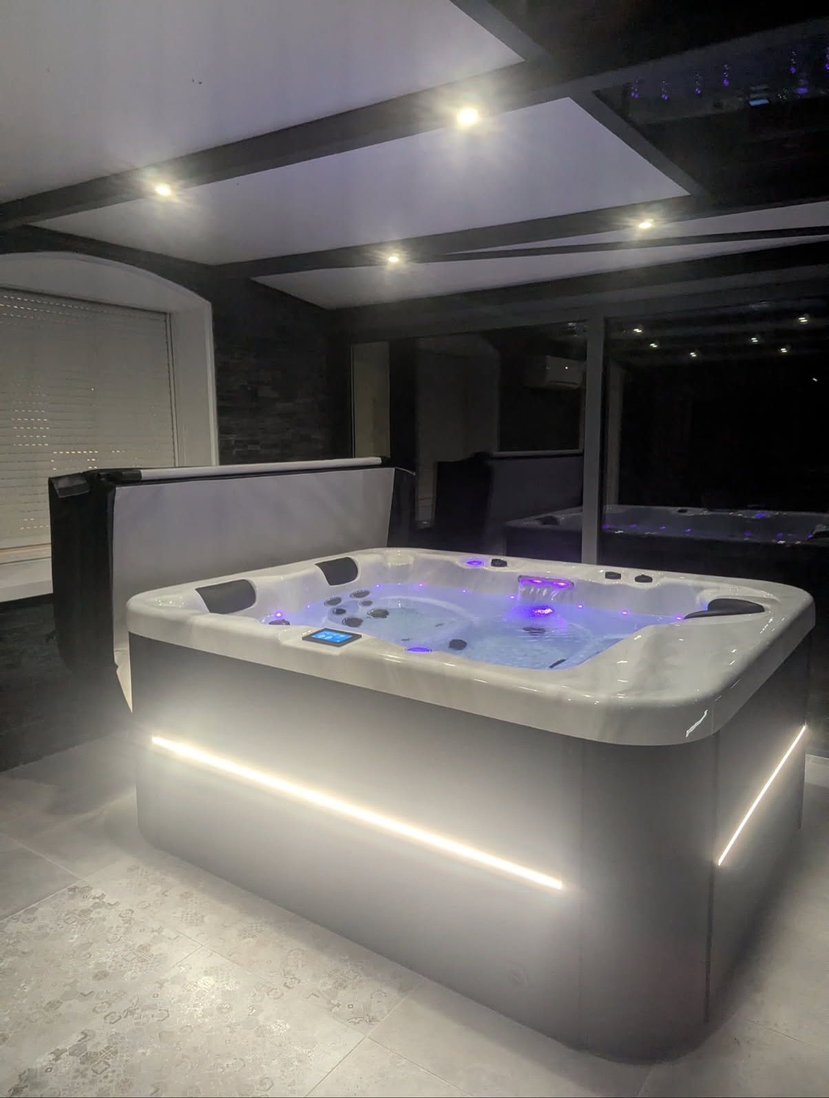 Un jacuzzi rectangulaire illuminé, dont l'eau est scintillante, est installé dans une pièce moderne dotée d'un éclairage encastré et de murs en pierre.