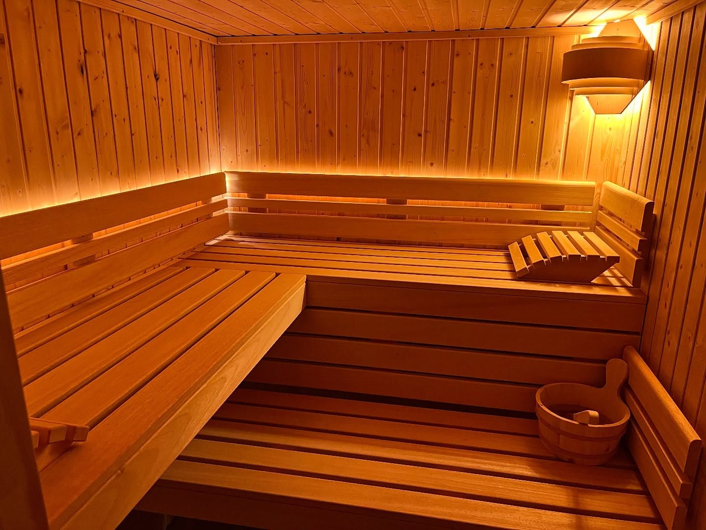 Vue intérieure d'un sauna en bois avec des banquettes étagées, un éclairage d'ambiance chaleureux et un seau en bois posé au sol.