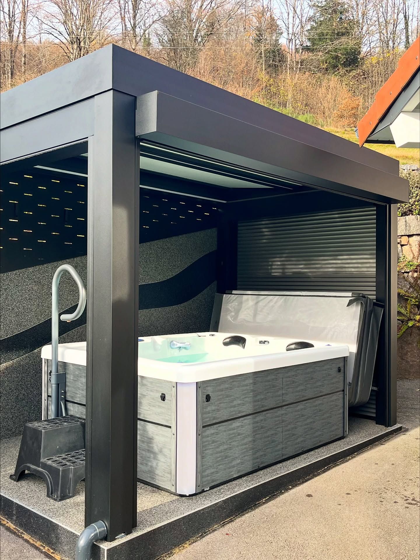 Un jacuzzi gris avec des marches assorties est installé sous une pergola moderne à persiennes noires, sur un sol en gravier à l'extérieur.
