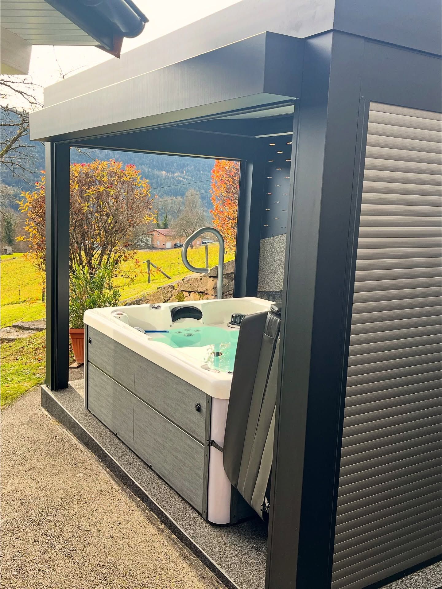 Une pergola moderne gris foncé abritant un jacuzzi blanc sur une terrasse en gravier, avec un paysage champêtre d'automne en arrière-plan.