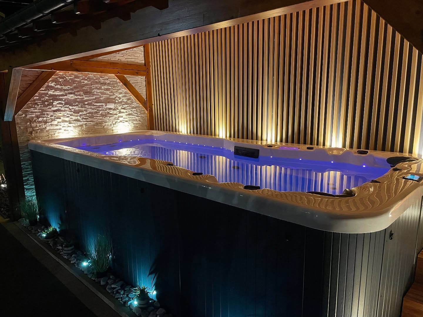 Un spa de nage rectangulaire éclairé en bleu se trouve sur un patio extérieur couvert aux murs en bois texturé et à l'éclairage d'ambiance chaleureux.