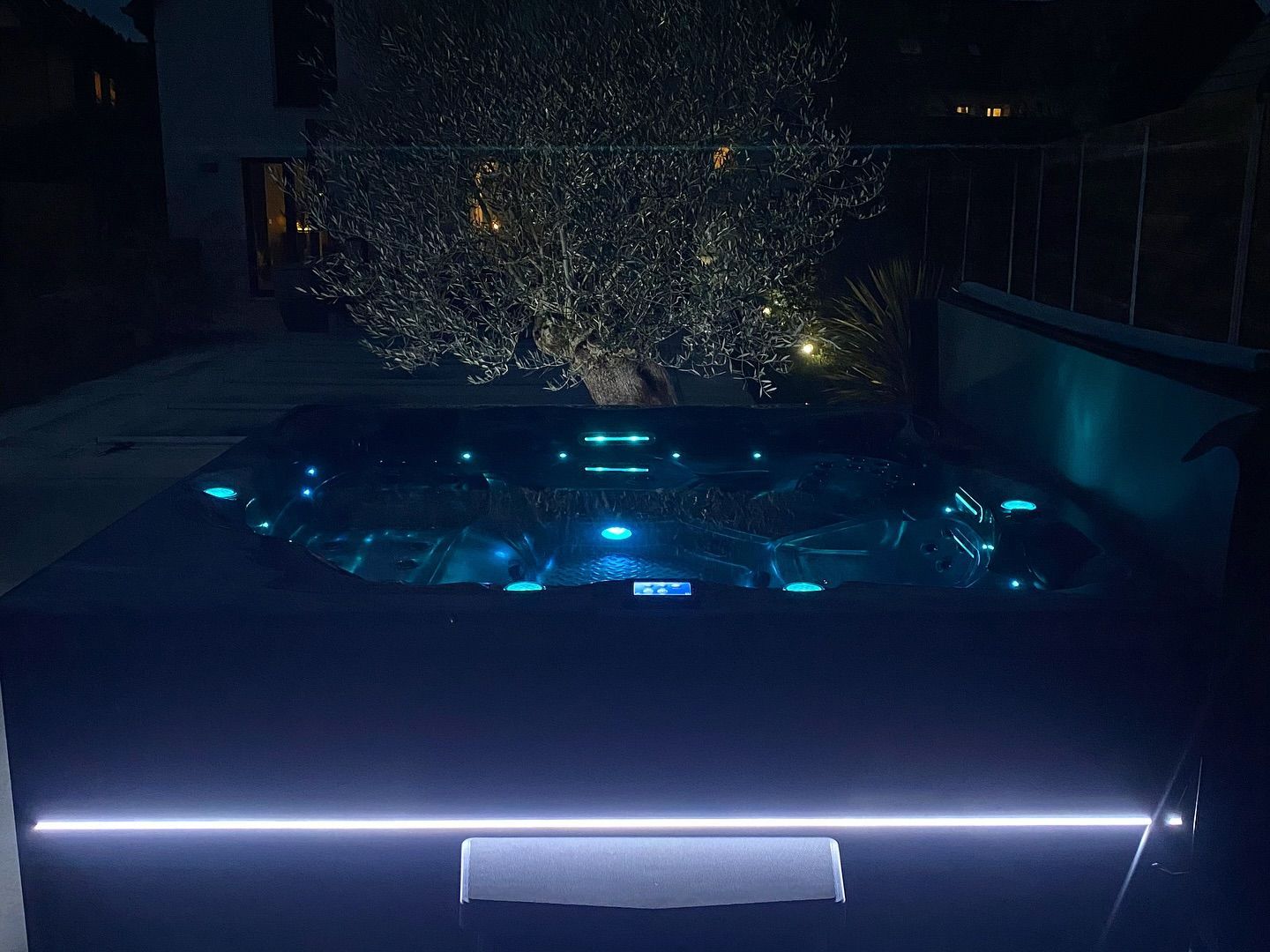 Un jacuzzi illuminé brille de lumières bleues à l'intérieur et d'une bande lumineuse blanche horizontale à l'extérieur la nuit.