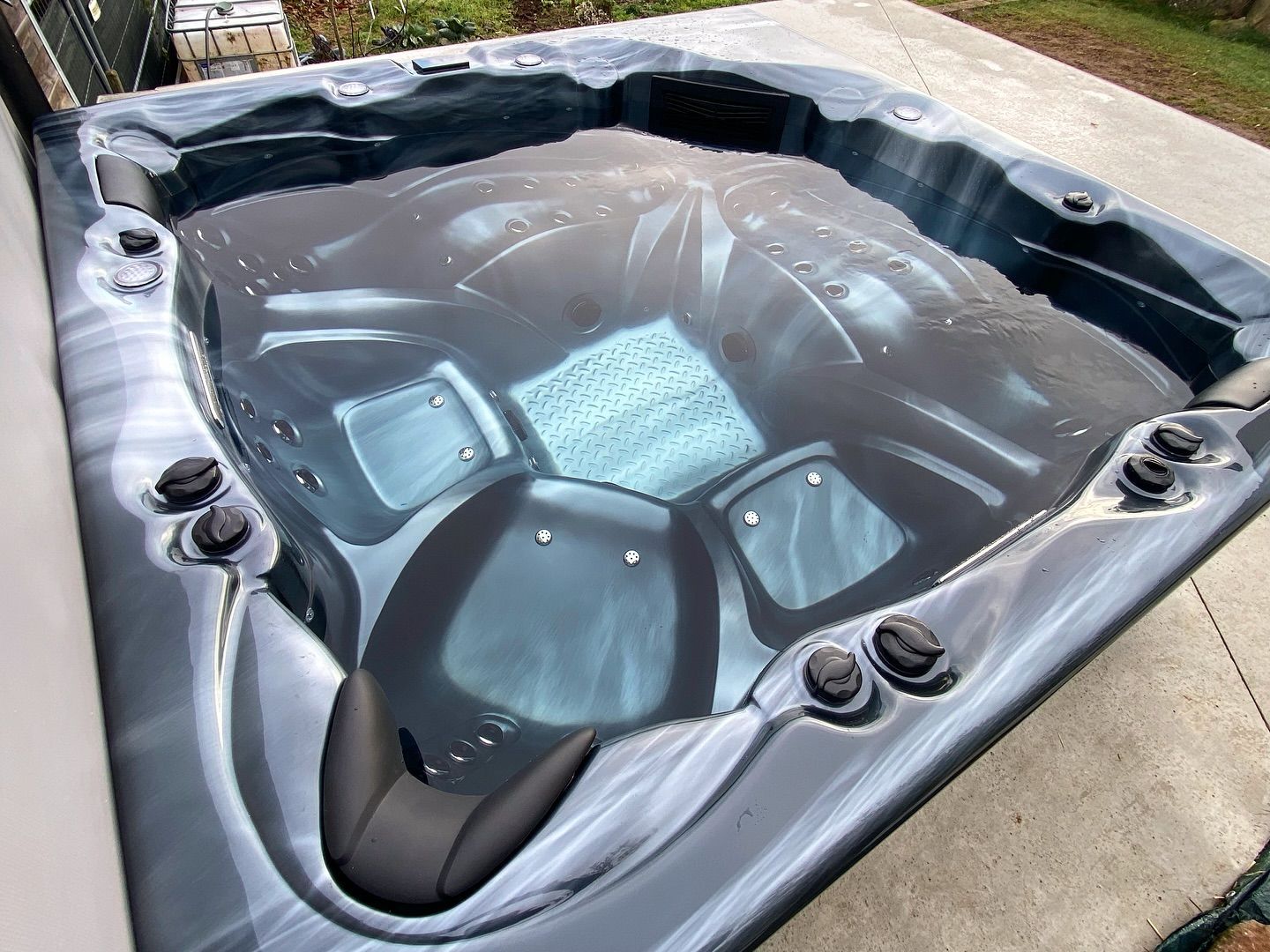Un jacuzzi sombre à la finition marbrée, rempli d'eau, avec des sièges moulés et des jets réglables, installé sur une terrasse en béton.