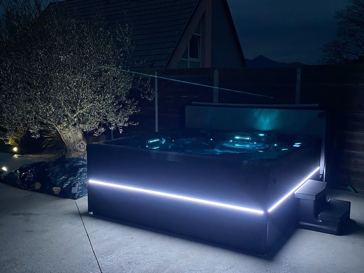 Un jacuzzi noir illuminé par des lumières intérieures bleues et une bande LED blanche le long de sa base, installé sur une terrasse la nuit.