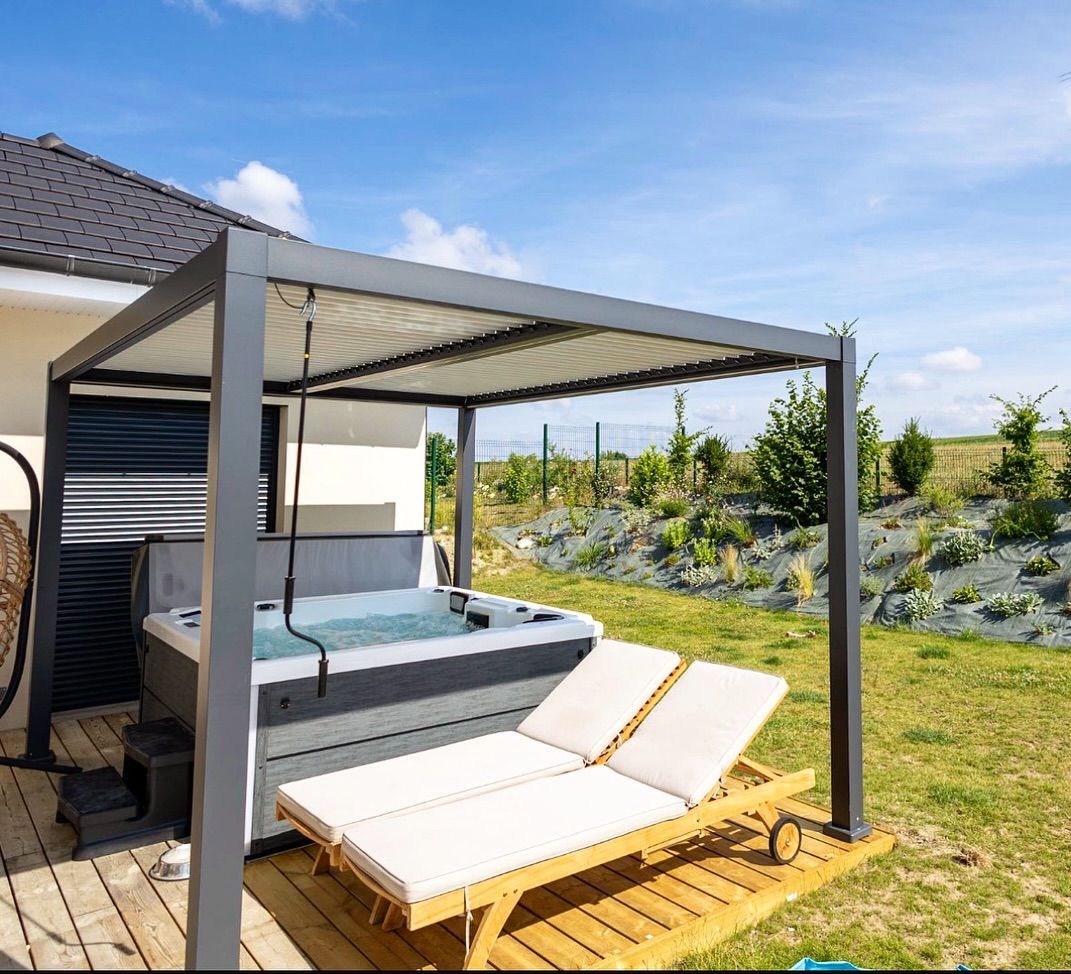 Un jacuzzi et deux chaises longues sont installés sur une terrasse en bois sous une pergola grise moderne, dans un jardin ensoleillé.