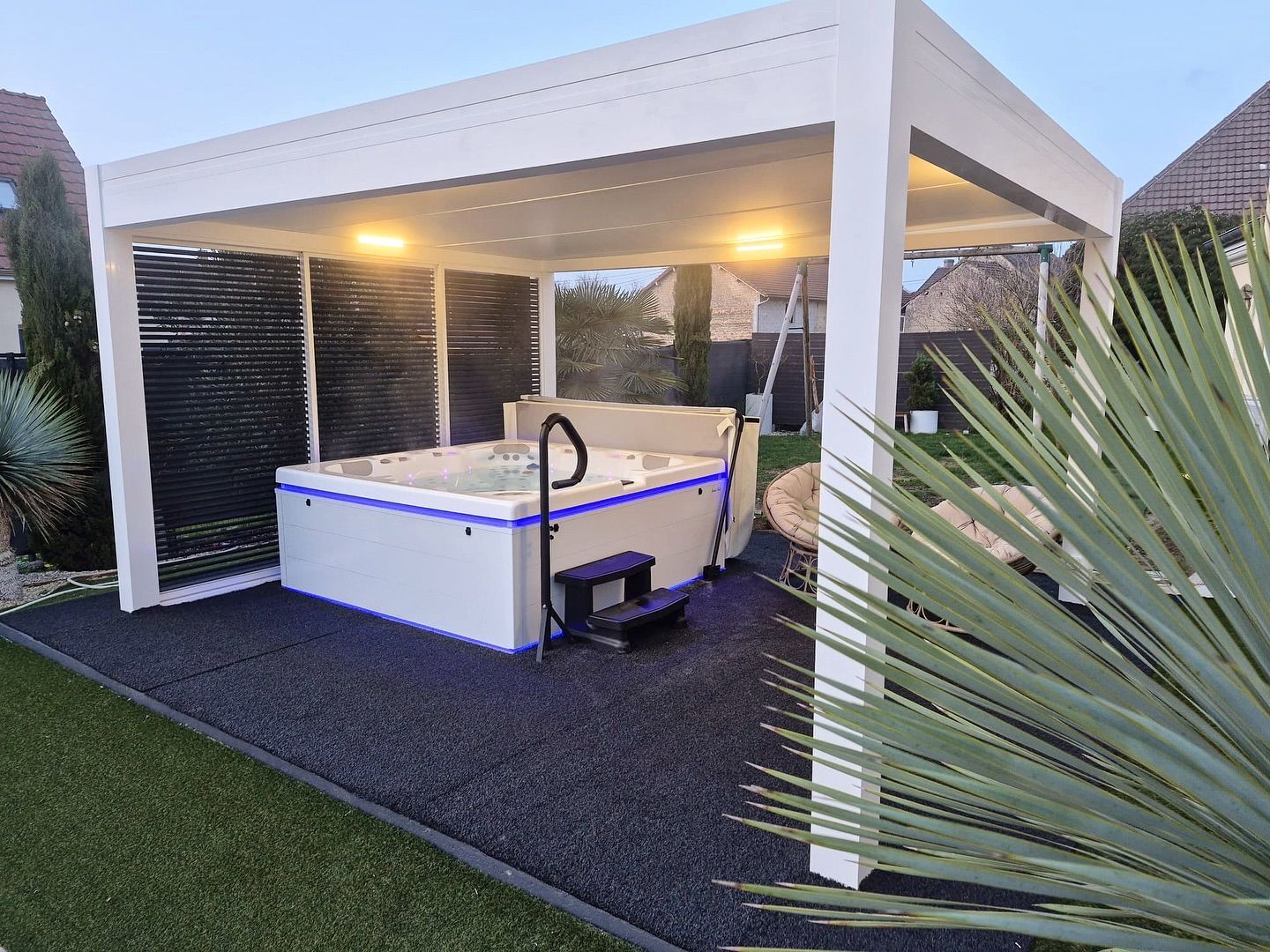 Un jacuzzi blanc avec éclairage LED bleu est installé sous une pergola moderne sur une terrasse en gravier foncé dans une cour arrière résidentielle.