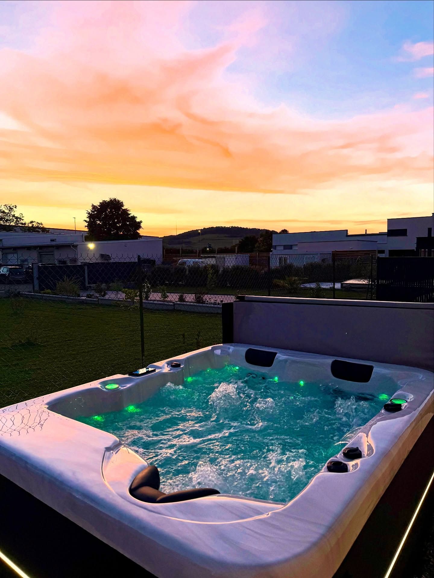 Un jacuzzi illuminé de lumières vertes scintillantes est installé à l'extérieur, sur une pelouse, au coucher du soleil, tandis que les teintes orangées et violettes flamboyantes illuminent la pièce.