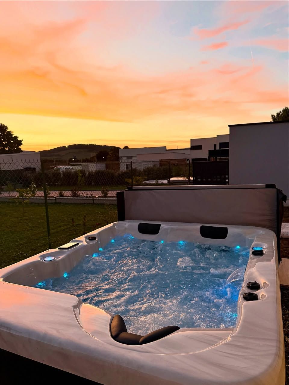 Au coucher du soleil, un jacuzzi illuminé se dresse dans une cour herbeuse, ses lumières bleues contrastant avec le ciel orangé.