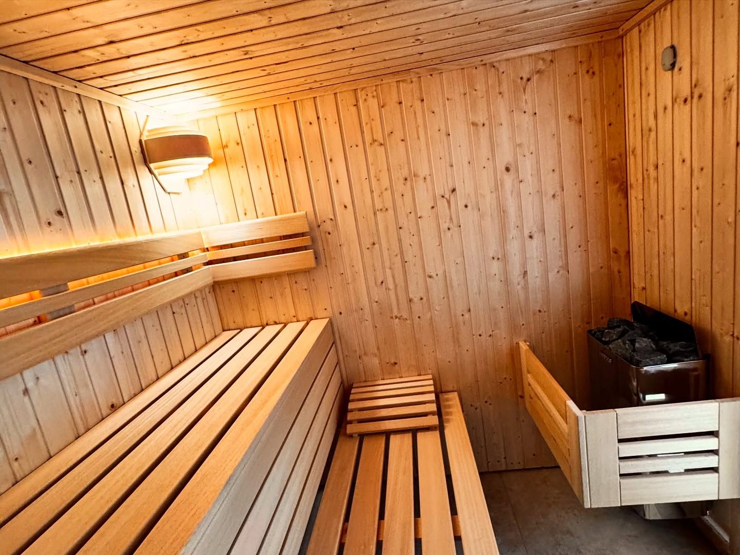 Intérieur d'un sauna en bois avec des banquettes étagées, un éclairage chaleureux et un poêle rempli de pierres sombres.