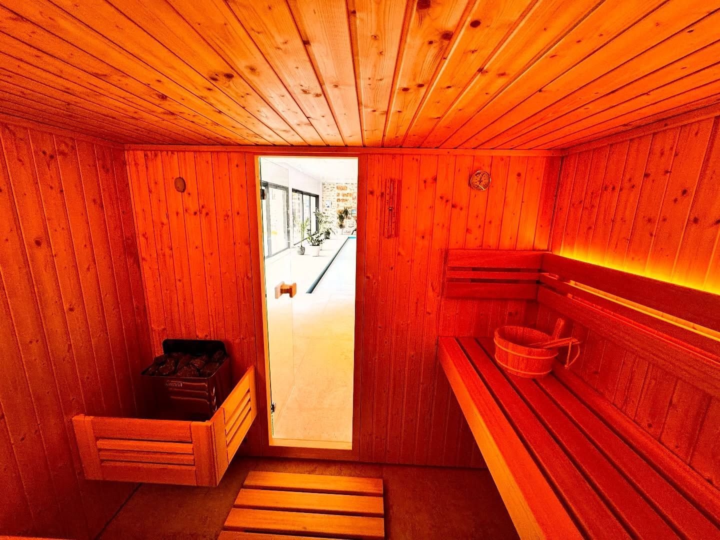 L'intérieur d'un sauna en bois comprend un poêle chauffant à gauche, un banc avec un seau à droite et une porte donnant sur un couloir.