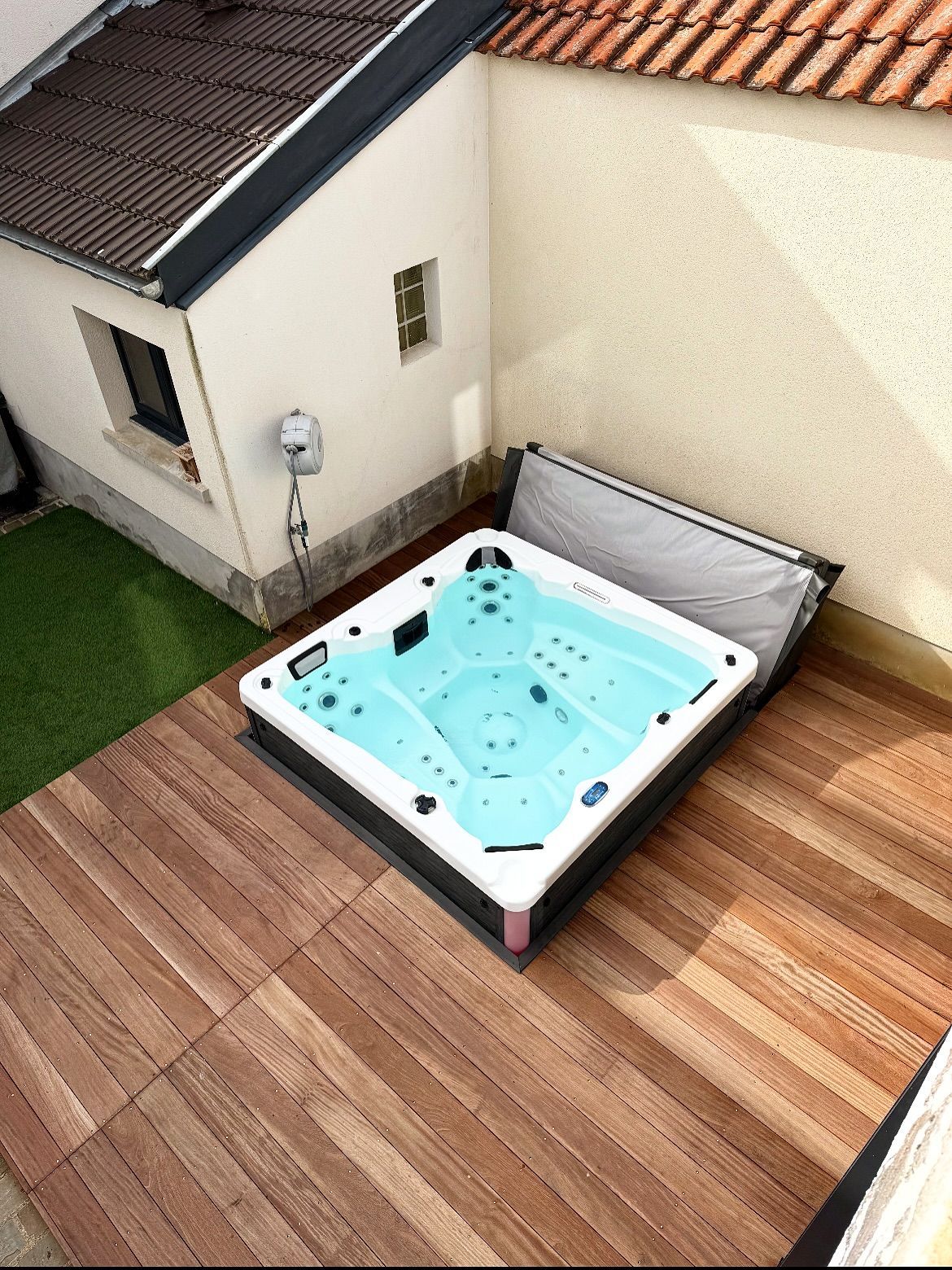 Un jacuzzi carré à l'eau bleu clair est installé sur une terrasse en bois, à côté d'un bâtiment blanc dont le toit est partiellement ouvert.