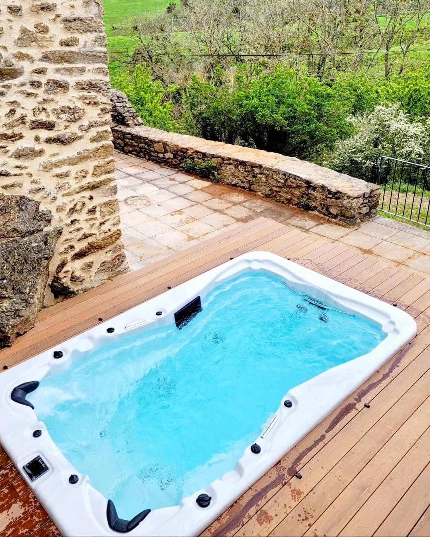 Un bain à remous est posé sur une terrasse en bois à côté d'un mur de pierre.
