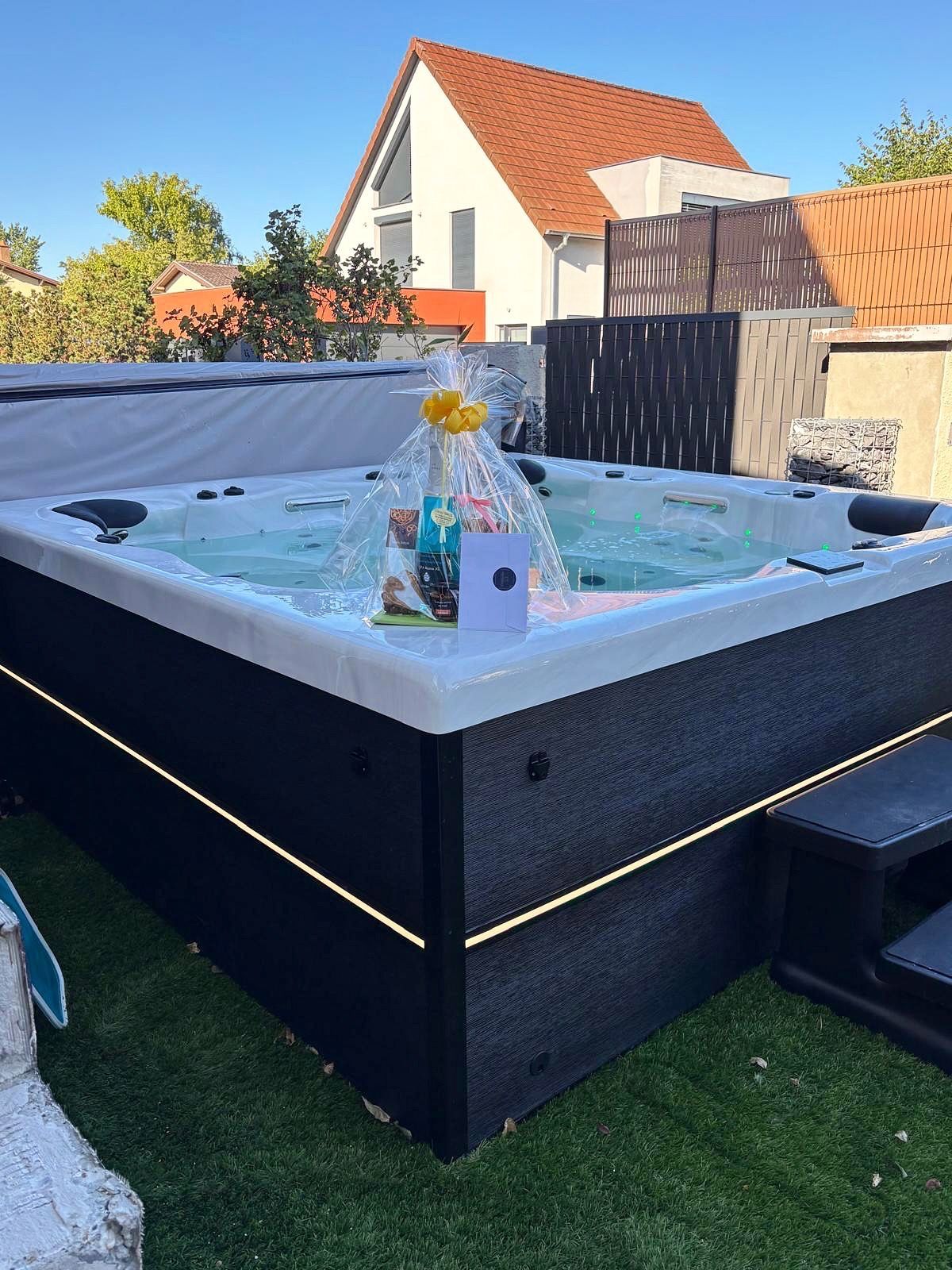 Jacuzzi extérieur avec champagne et panier-cadeau. Extérieur noir, bordures éclairées, eau bouillonnante.