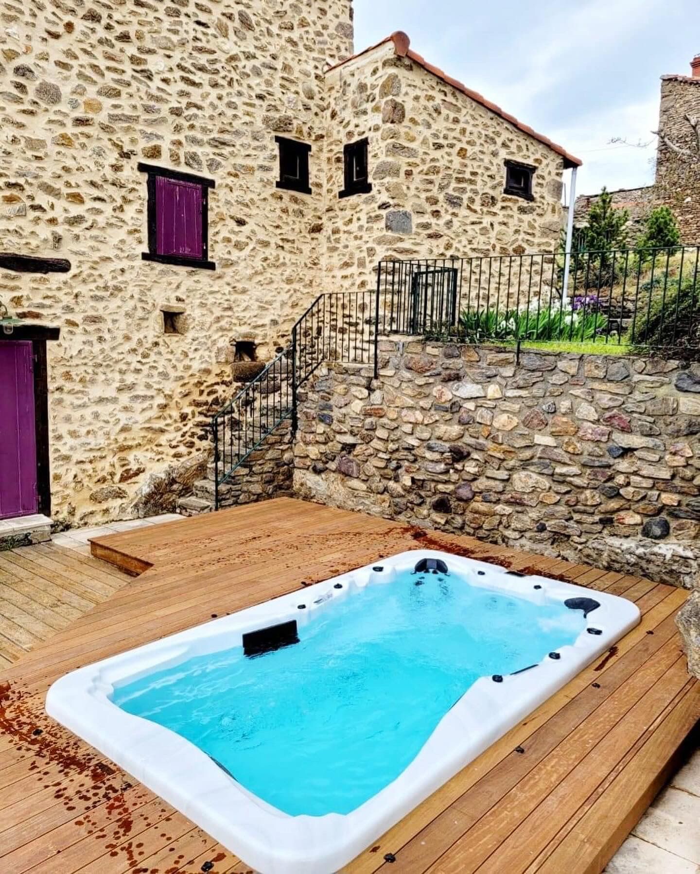 Un bain à remous est installé sur une terrasse en bois devant un bâtiment en pierre.