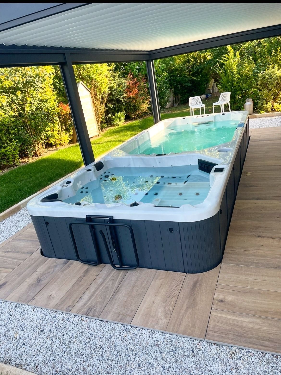 Spa de nage sous une pergola, installé sur des dalles de bois. Le spa dispose d'un jacuzzi et d'un espace de nage, à proximité de deux chaises blanches.