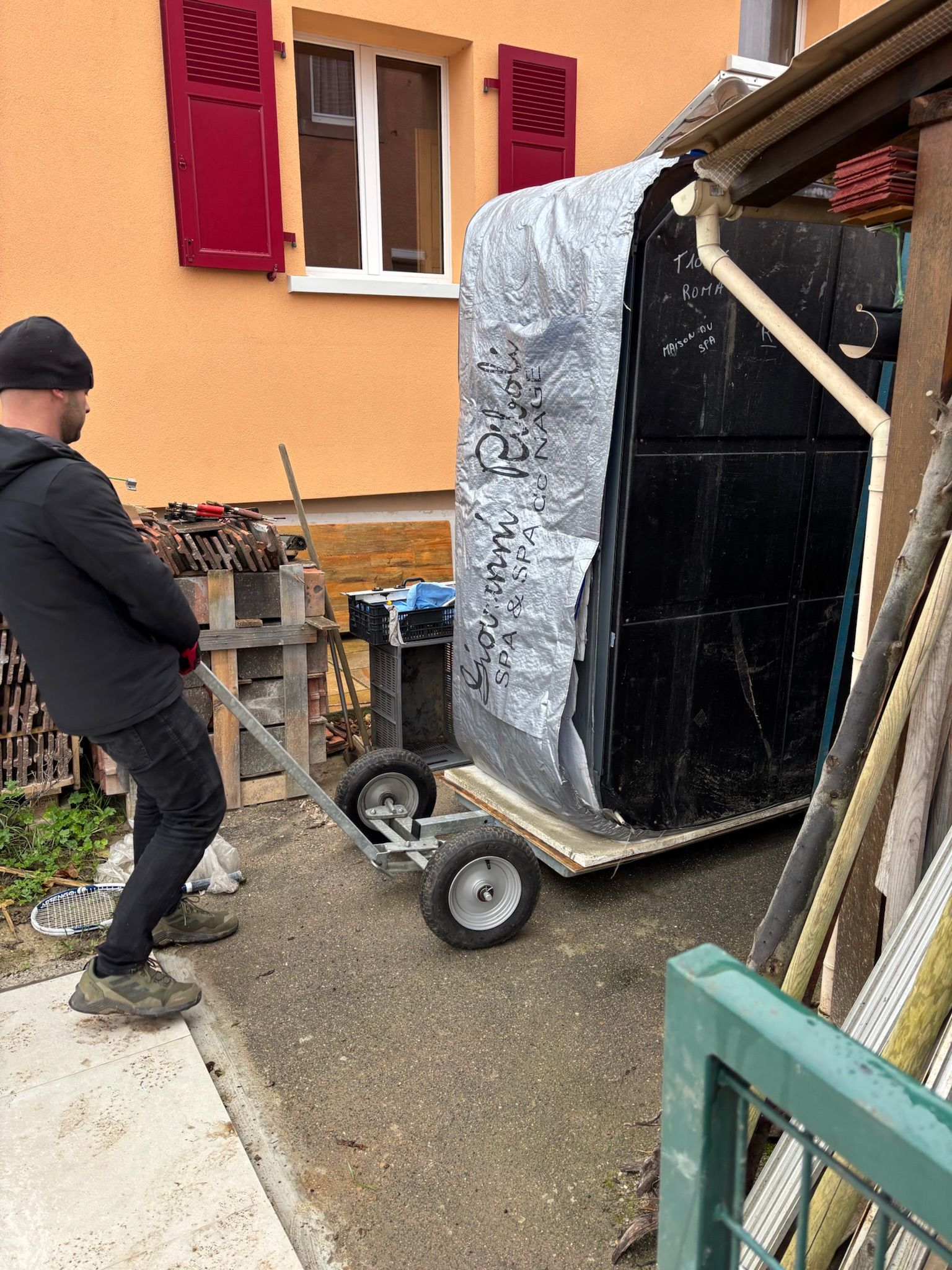 Un homme pousse un chariot avec un gros objet dessus devant un bâtiment.
