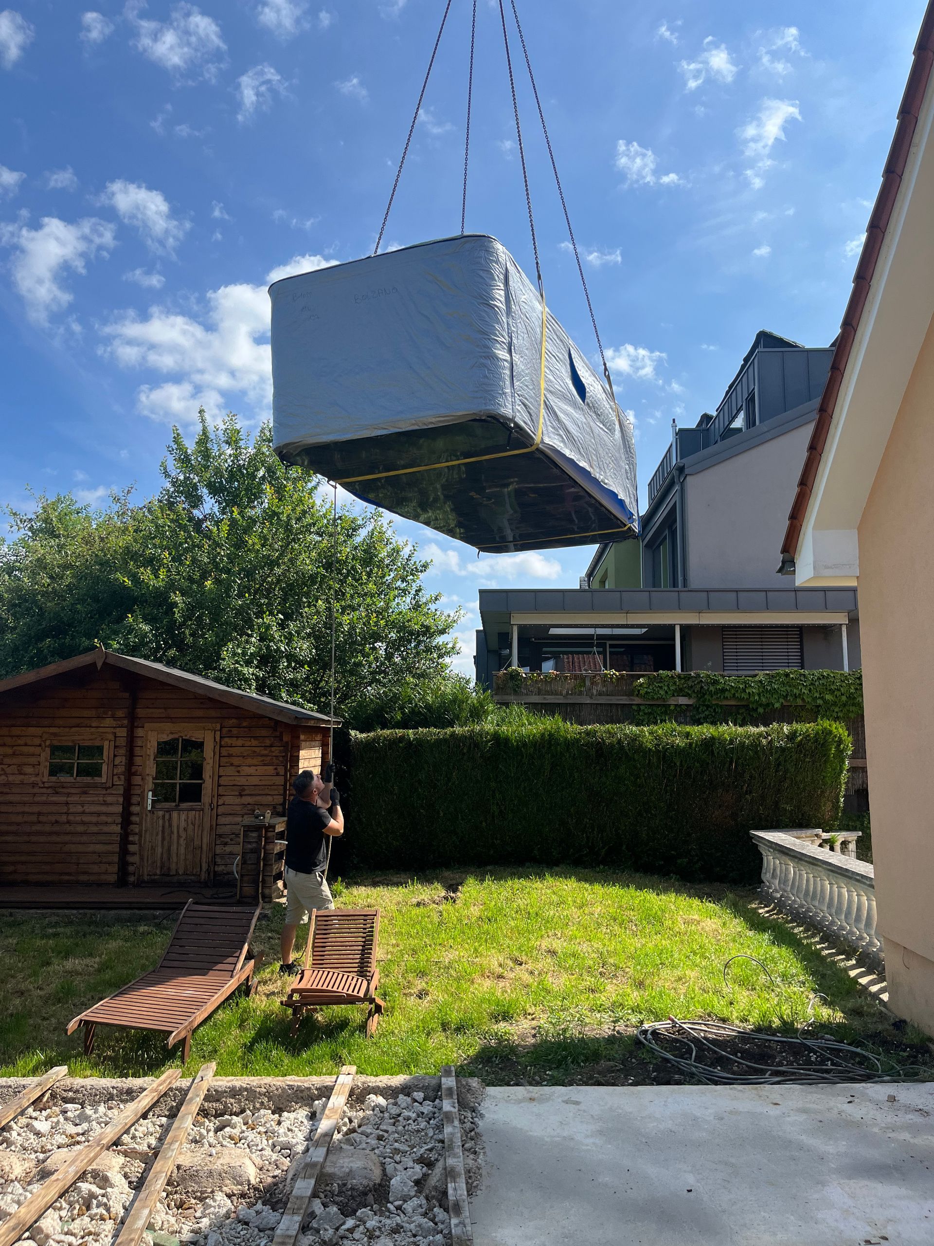 Une grande boîte blanche est soulevée par une grue dans une arrière-cour.