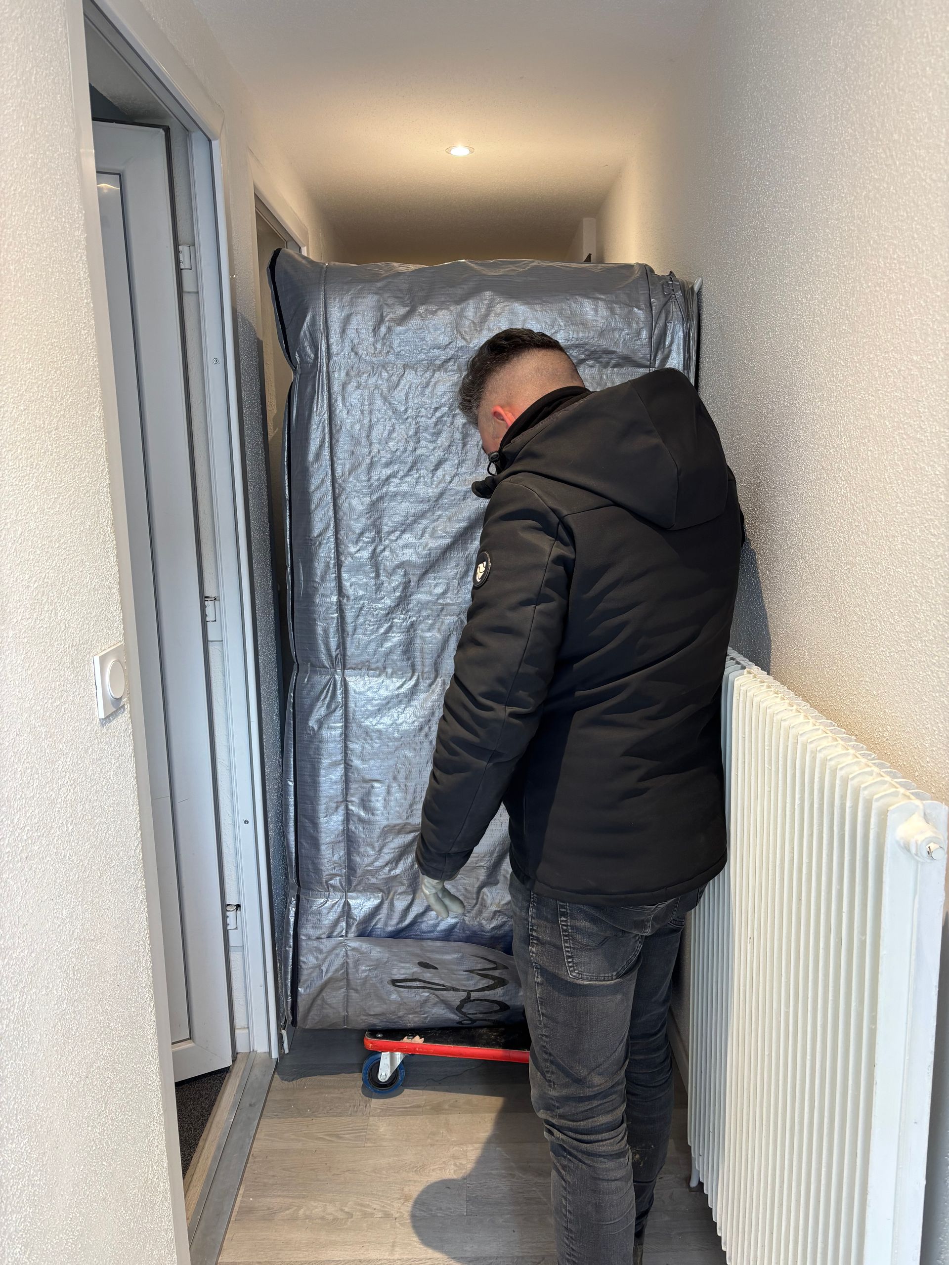Un homme en veste noire déplace un gros meuble dans un couloir.