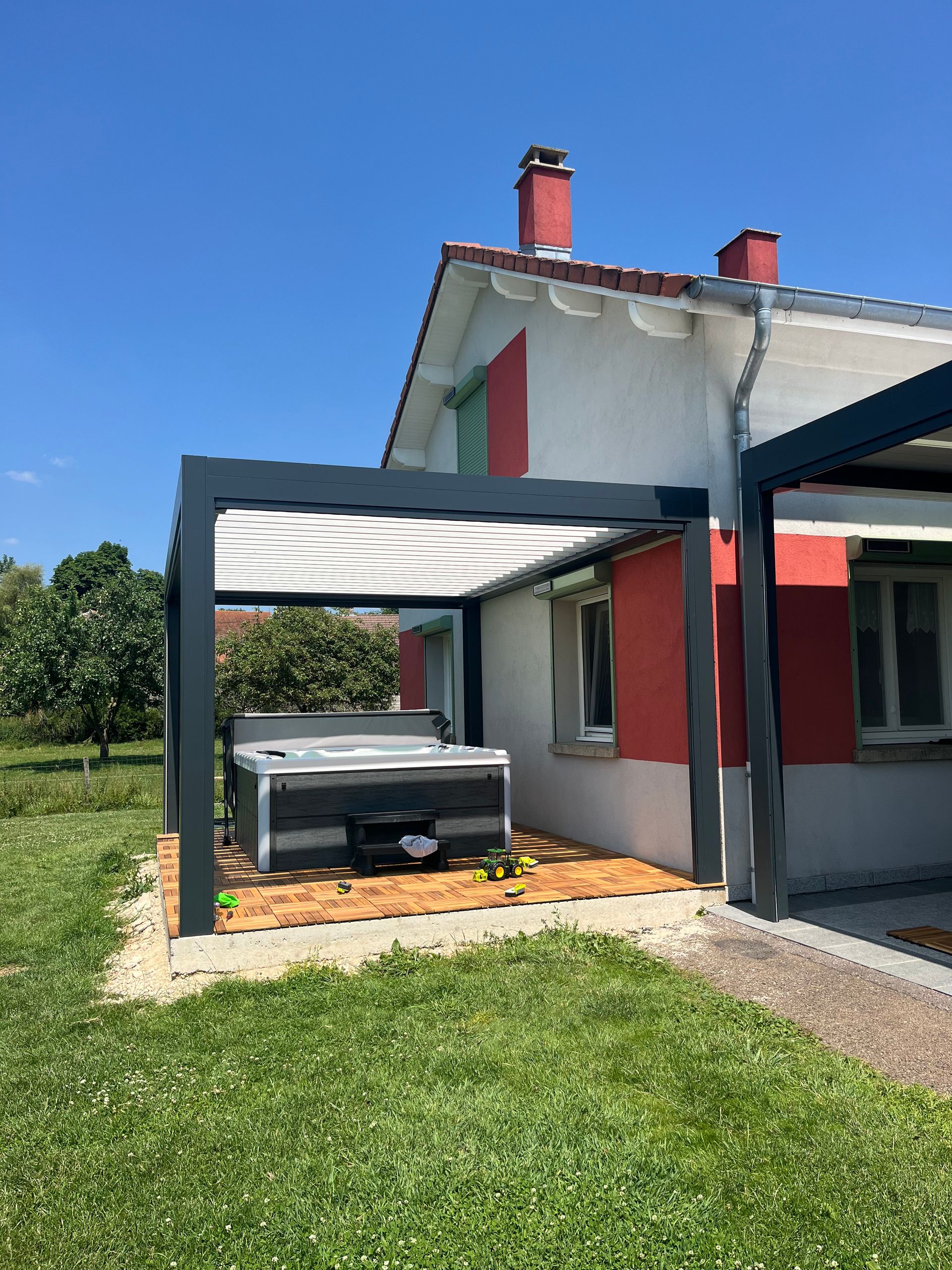 Un jacuzzi est installé sous une pergola devant une maison.