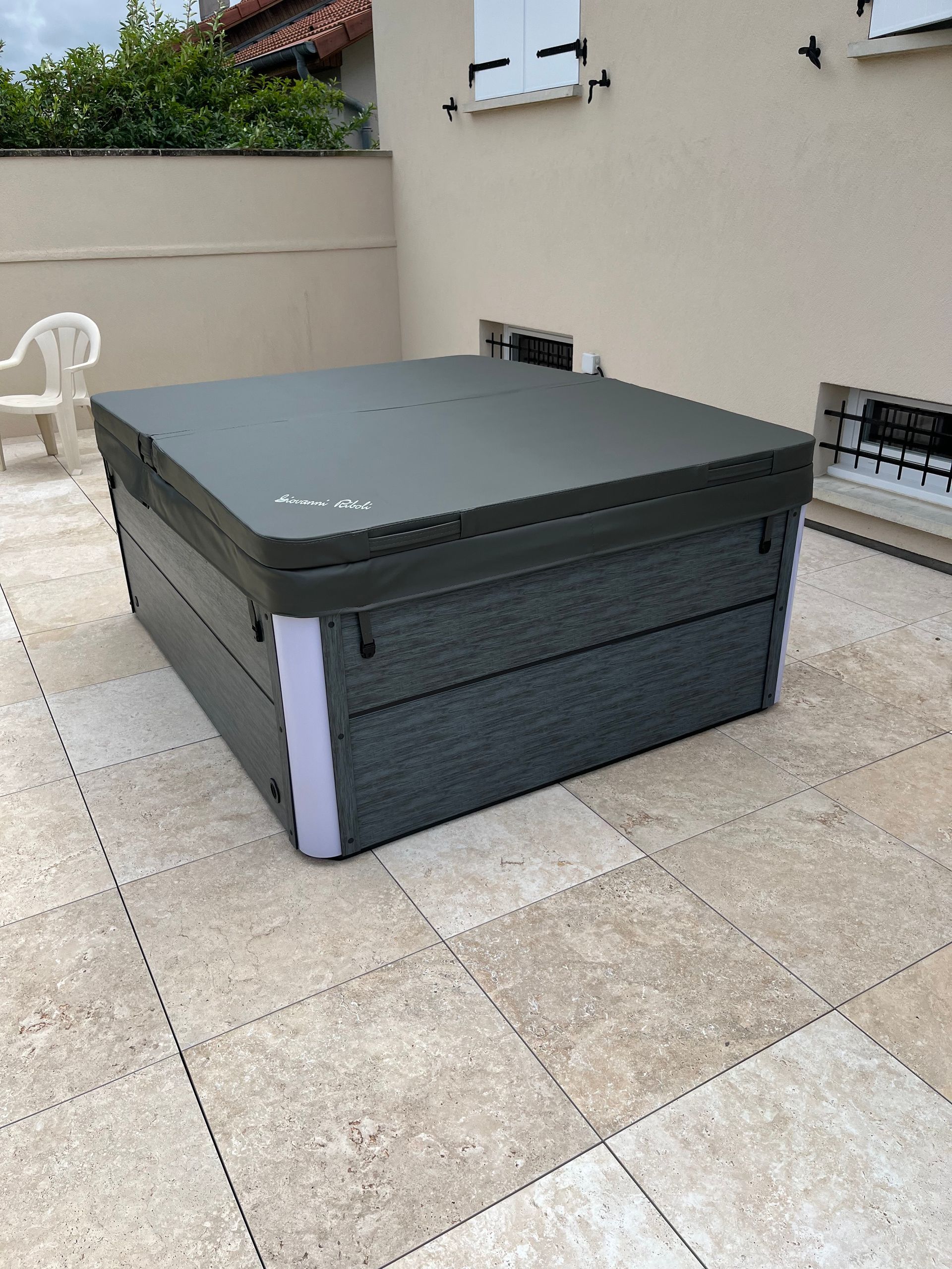 Spa carré avec couverture gris foncé sur un patio en pierre, avec des murs blancs et beiges et du feuillage en arrière-plan.