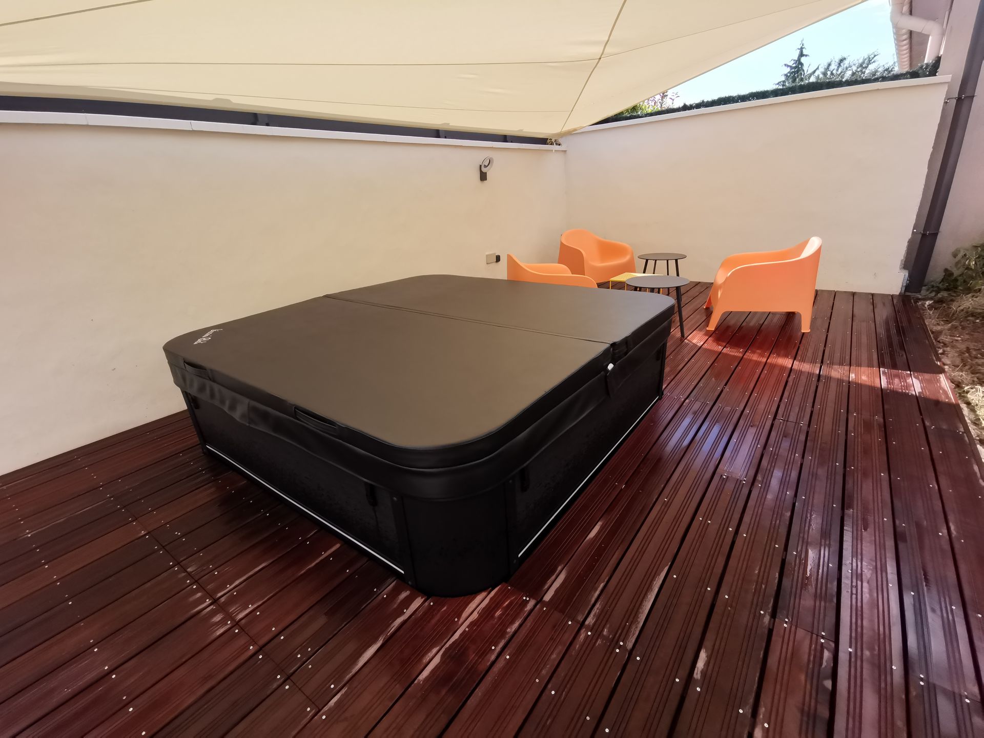 Un jacuzzi noir, recouvert de sa couverture, est installé sur une terrasse en bois sombre, sous une voile d'ombrage beige, avec des chaises orange à proximité.