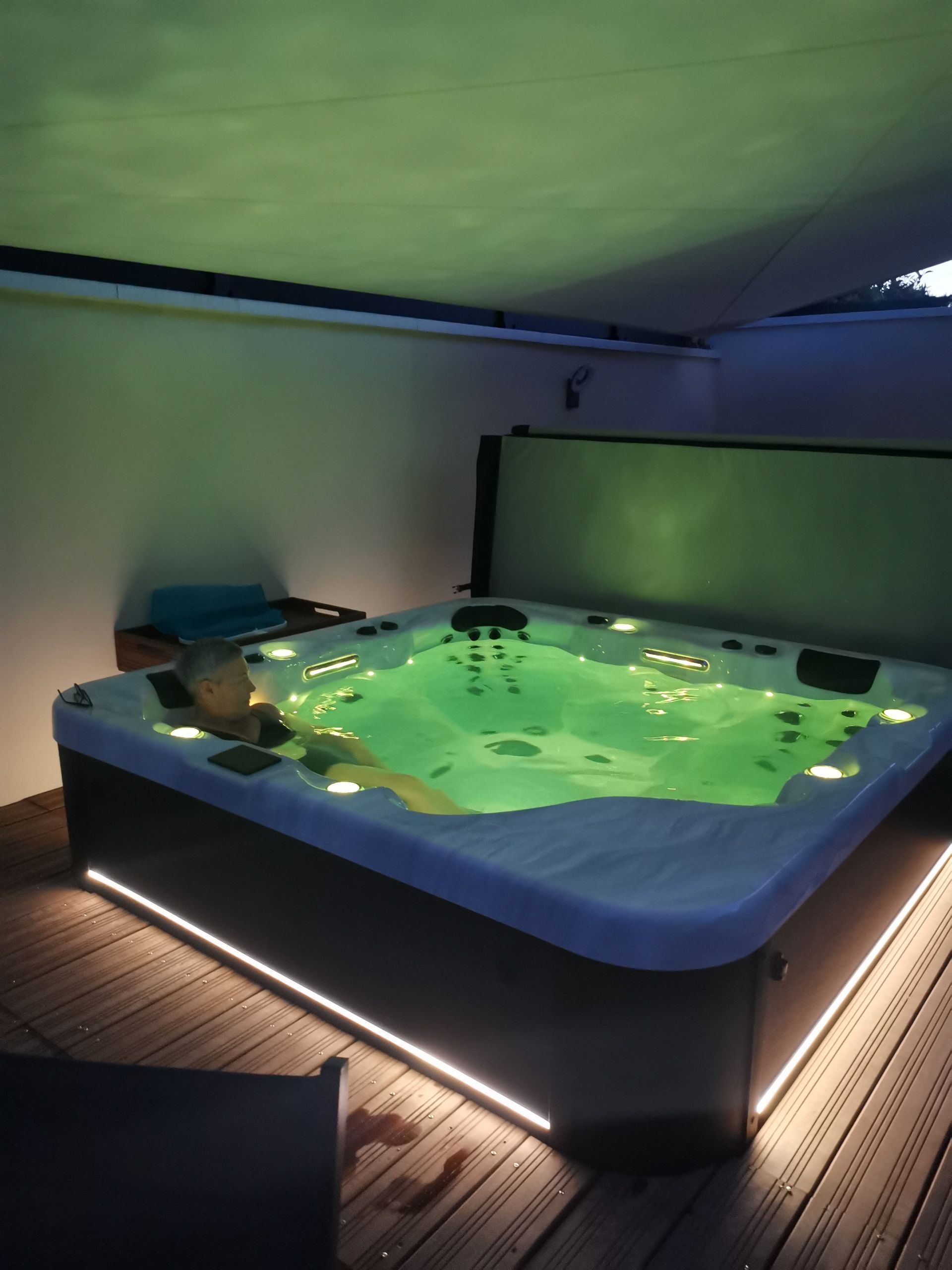 Une personne se détend dans un bain à remous extérieur d'un vert éclatant, installé sur une terrasse en bois sous un auvent ombragé, la nuit.