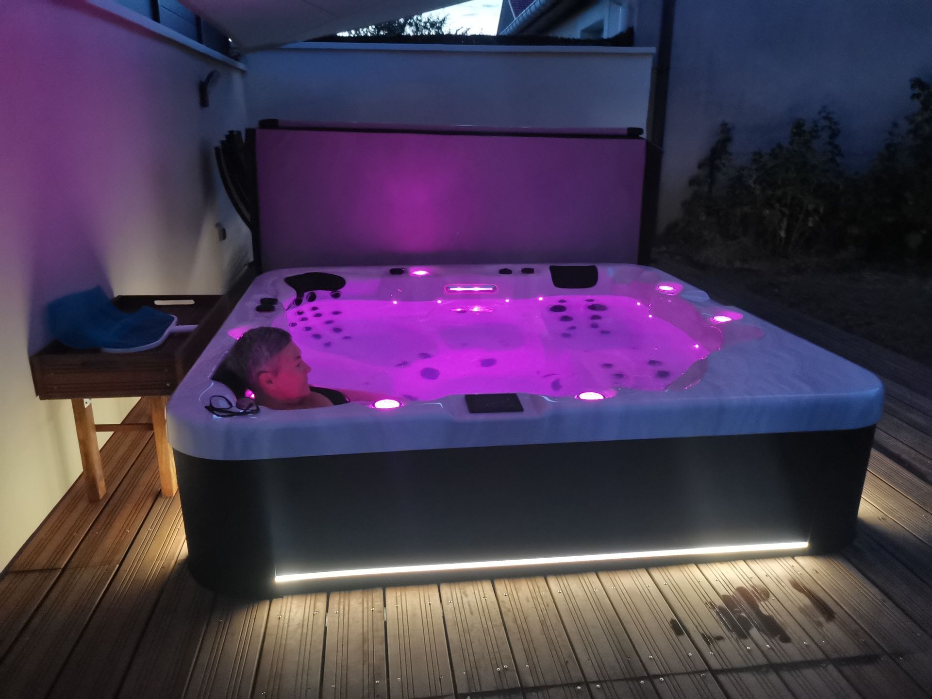 Une personne se détend de nuit dans un jacuzzi illuminé d'une douce lumière violette, installé sur une terrasse en bois, avec une couverture ouverte derrière elle.