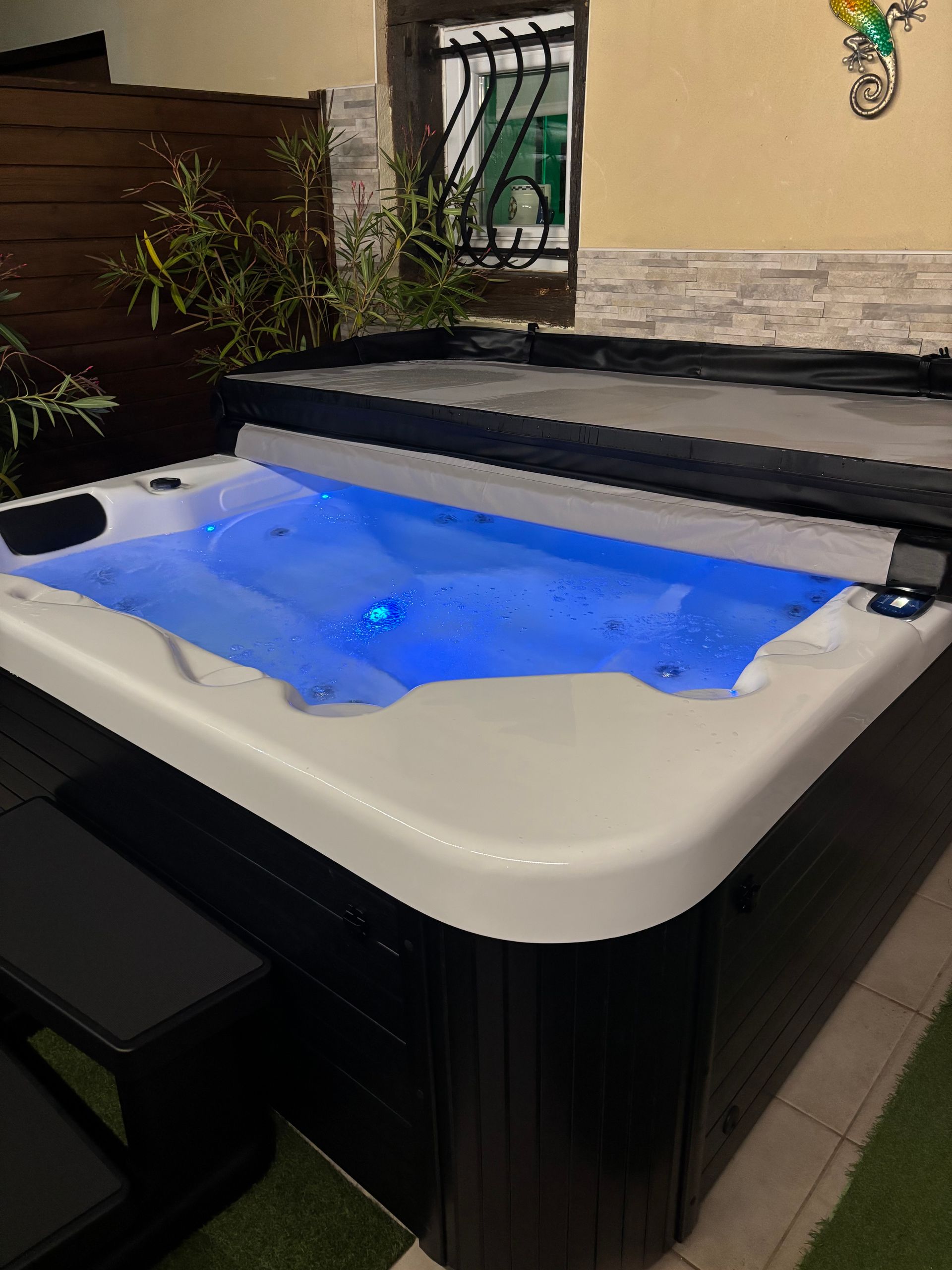 Jacuzzi éclairé avec eau bleue, couvercle ouvert, près d'une clôture en bois et d'une fenêtre.