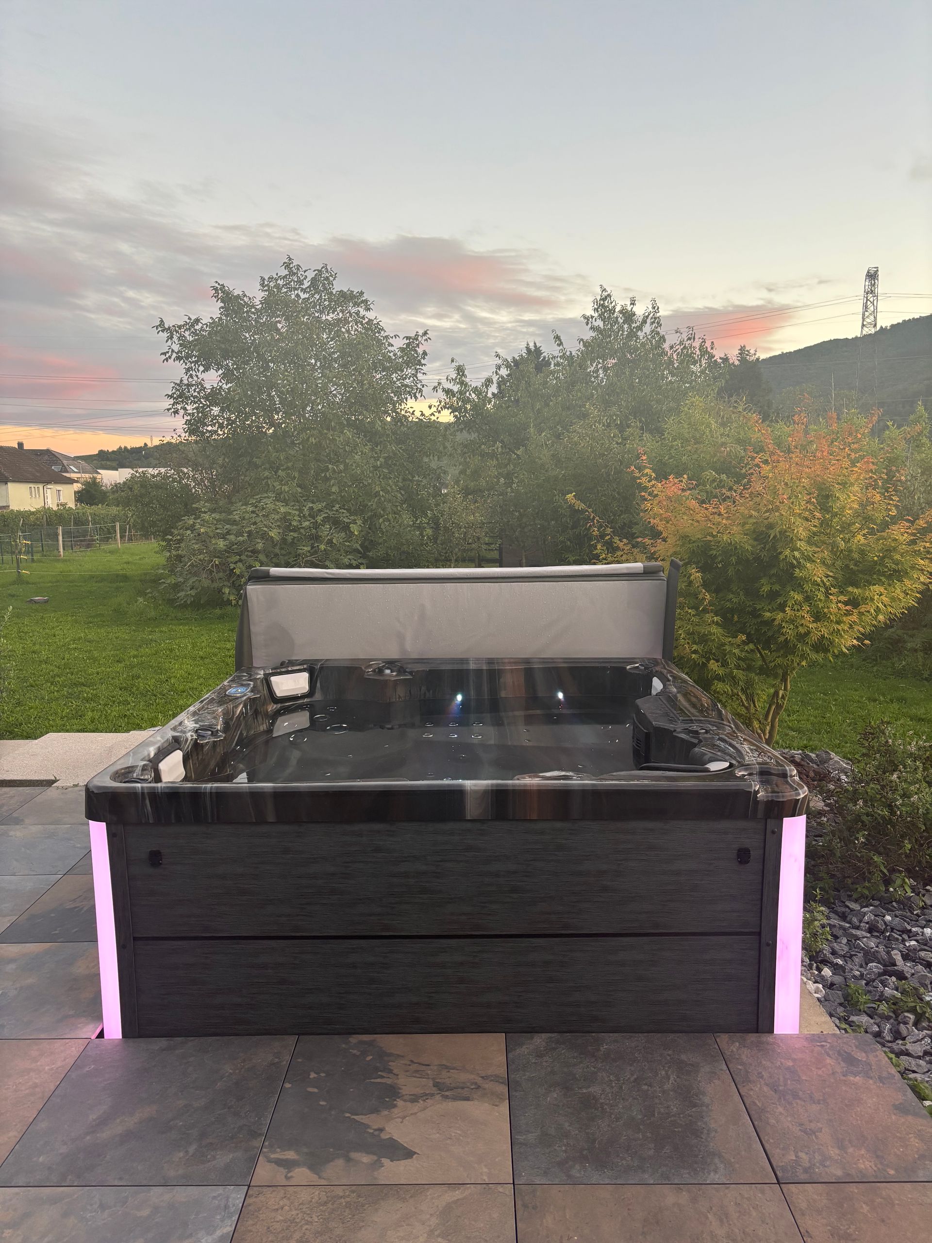 Bain à remous avec lumières roses, installé sur une terrasse au crépuscule, avec des arbres et une colline en arrière-plan.