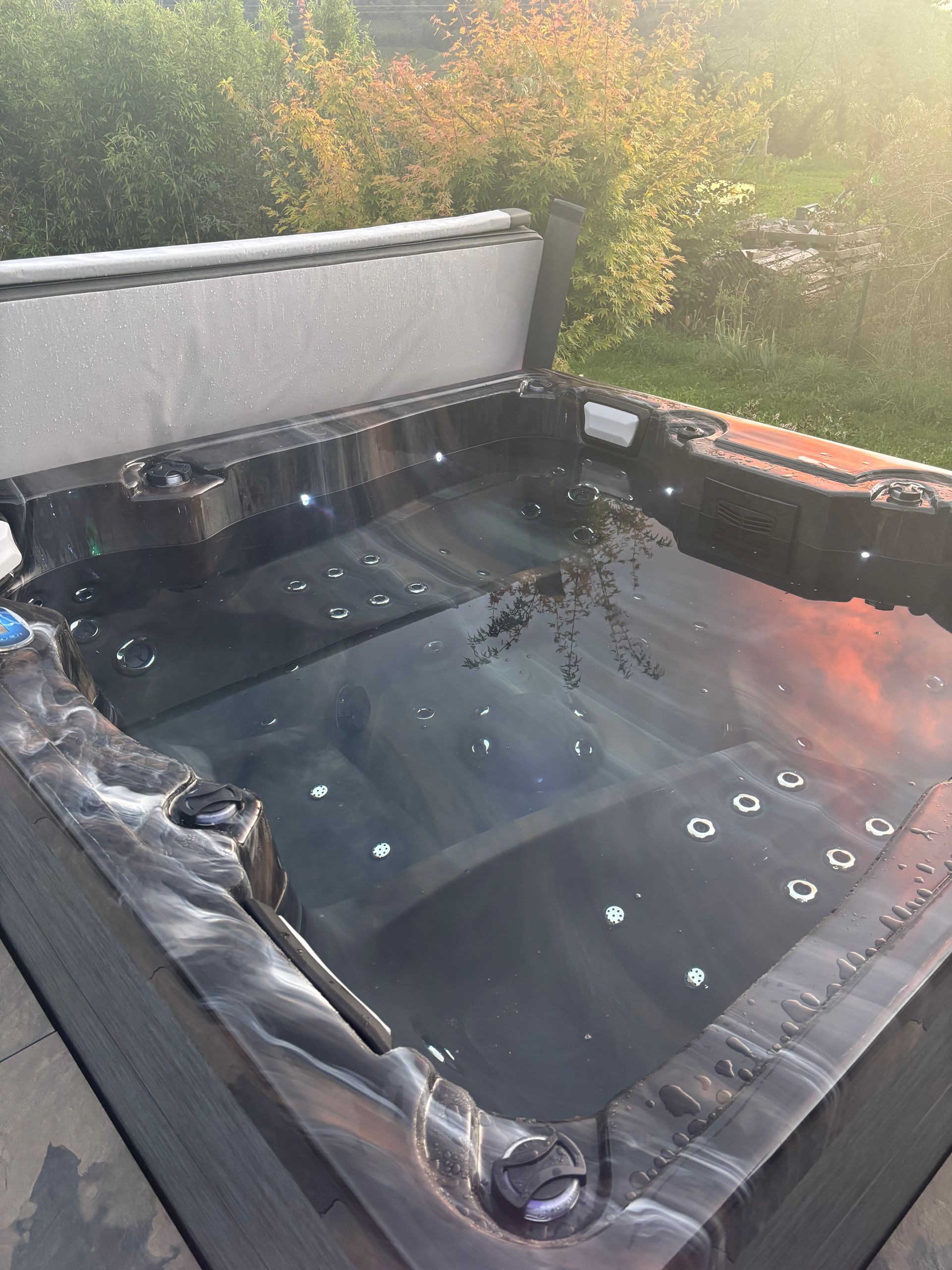Jacuzzi rempli d'eau, entouré d'une terrasse gris foncé, couvercle ouvert. Fond vert luxuriant.