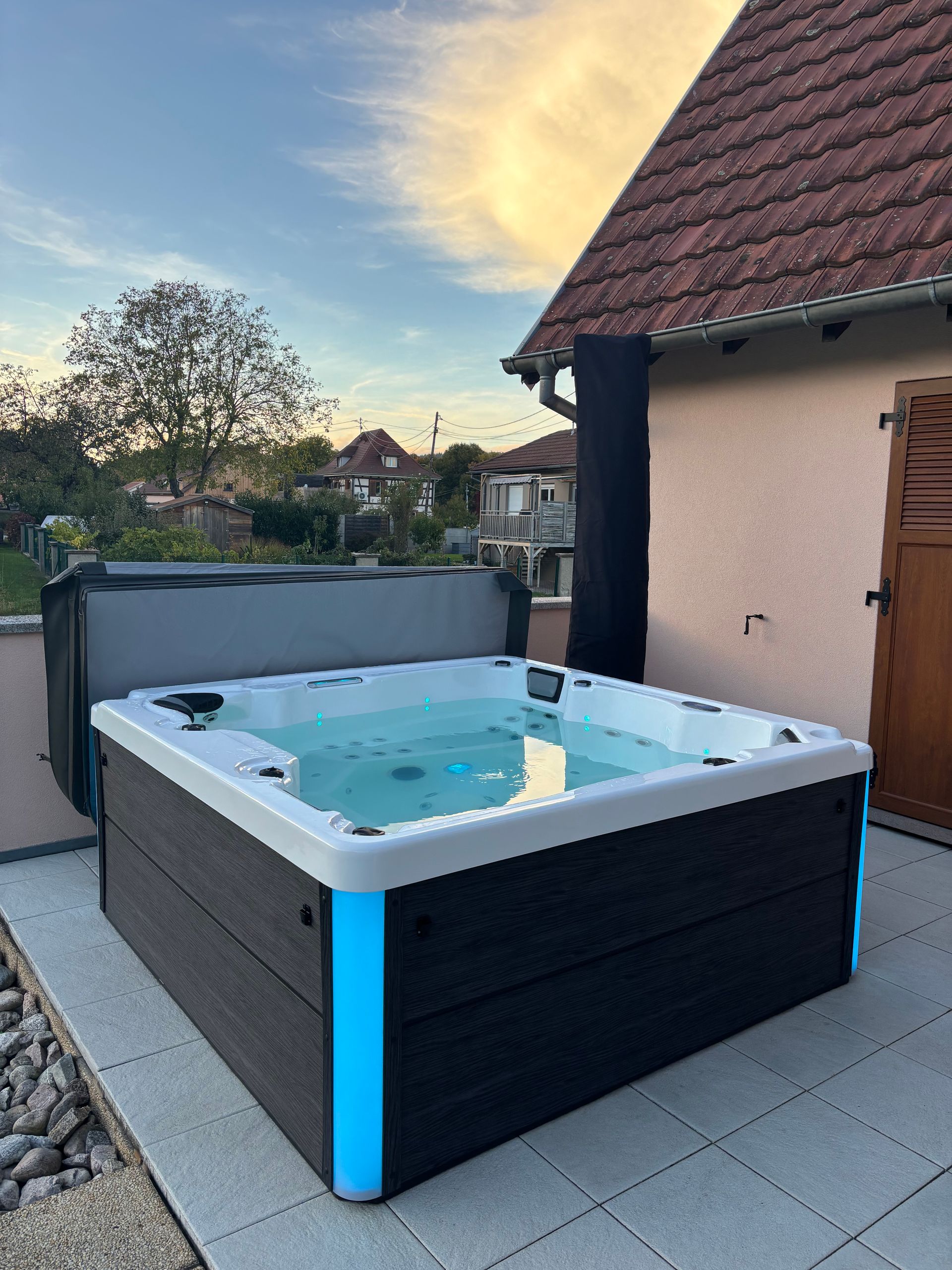 Un jacuzzi carré avec des lumières bleues se trouve sur une terrasse, partiellement recouvert de son couvercle gris.